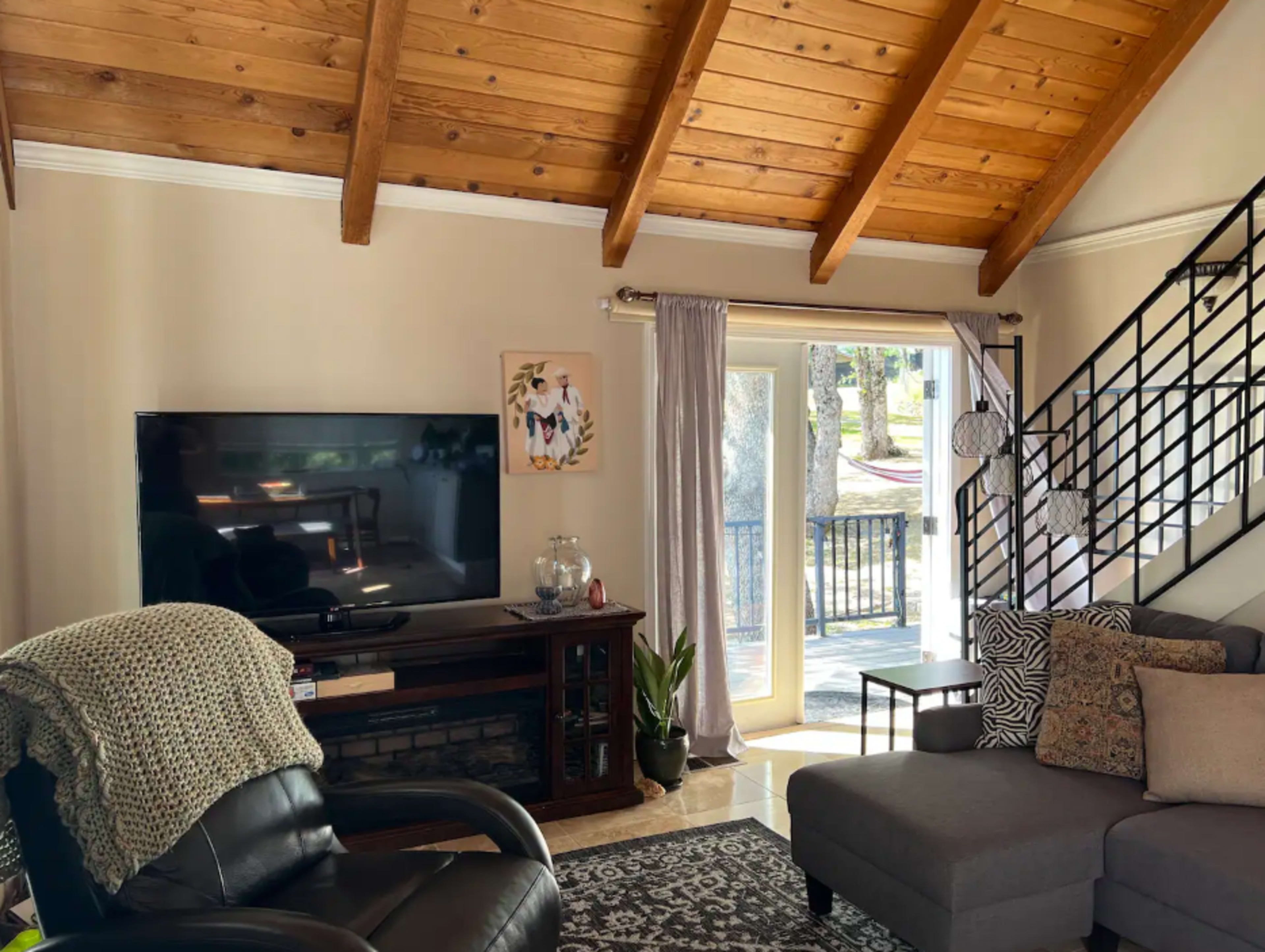 A spacious living room features a large television, an L-shaped couch, and a door leading to an outdoor area, all highlighted by wooden beams on the ceiling.