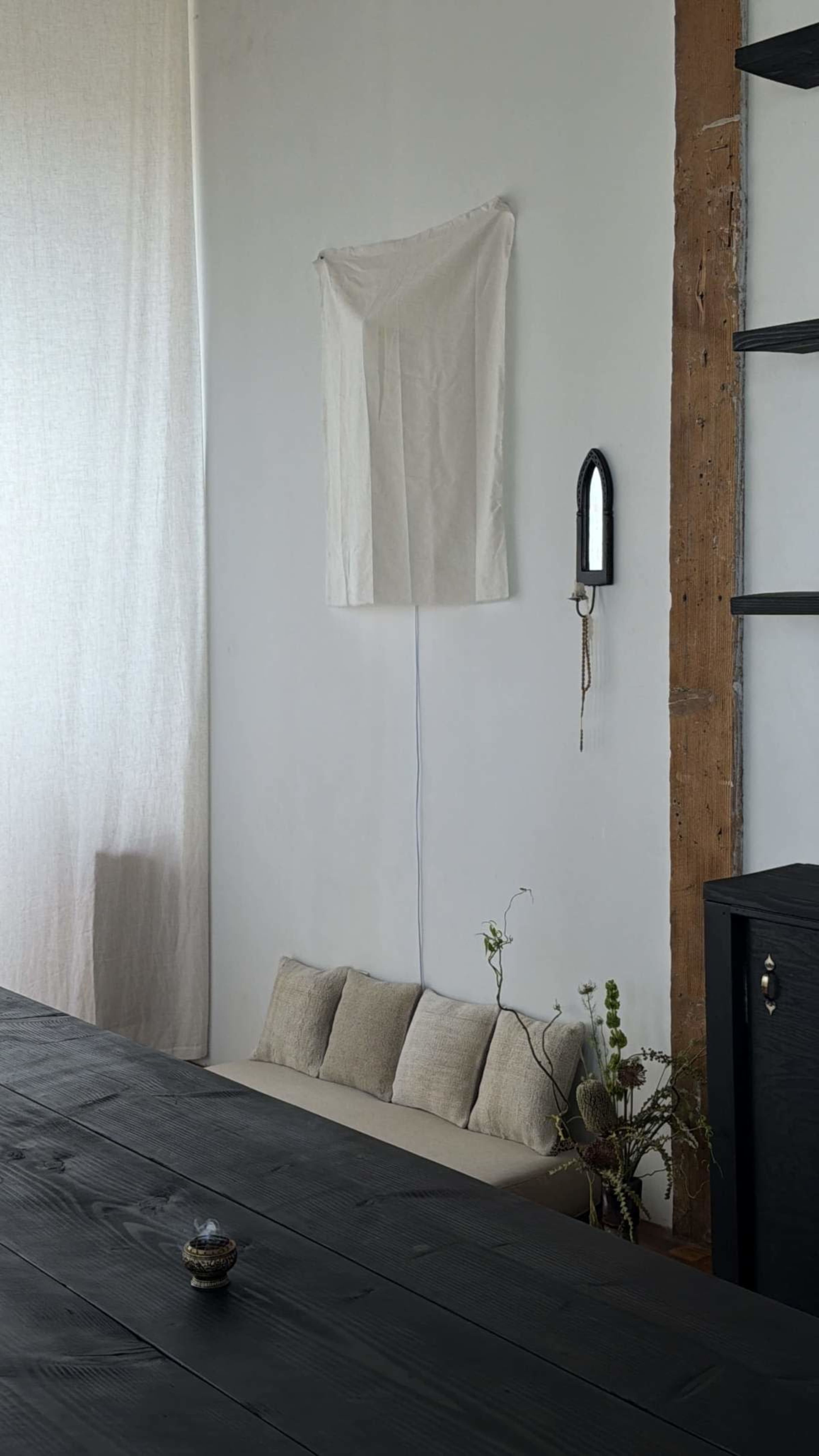 A simple interior scene features a wooden table, a wall with a hanging fabric piece, and a small mirror mounted nearby, with cushions placed on a ledge and a plant in the corner.