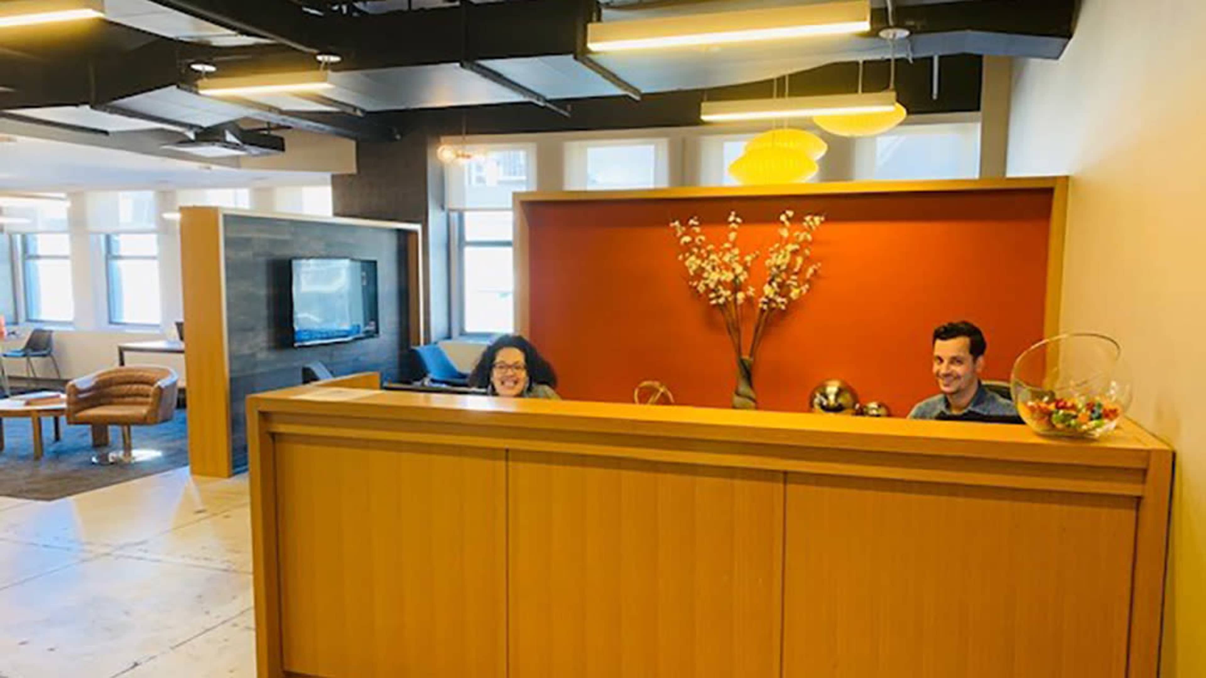 Two receptionists are seated behind a wooden front desk in a modern office space with bright lighting and a welcoming atmosphere.
