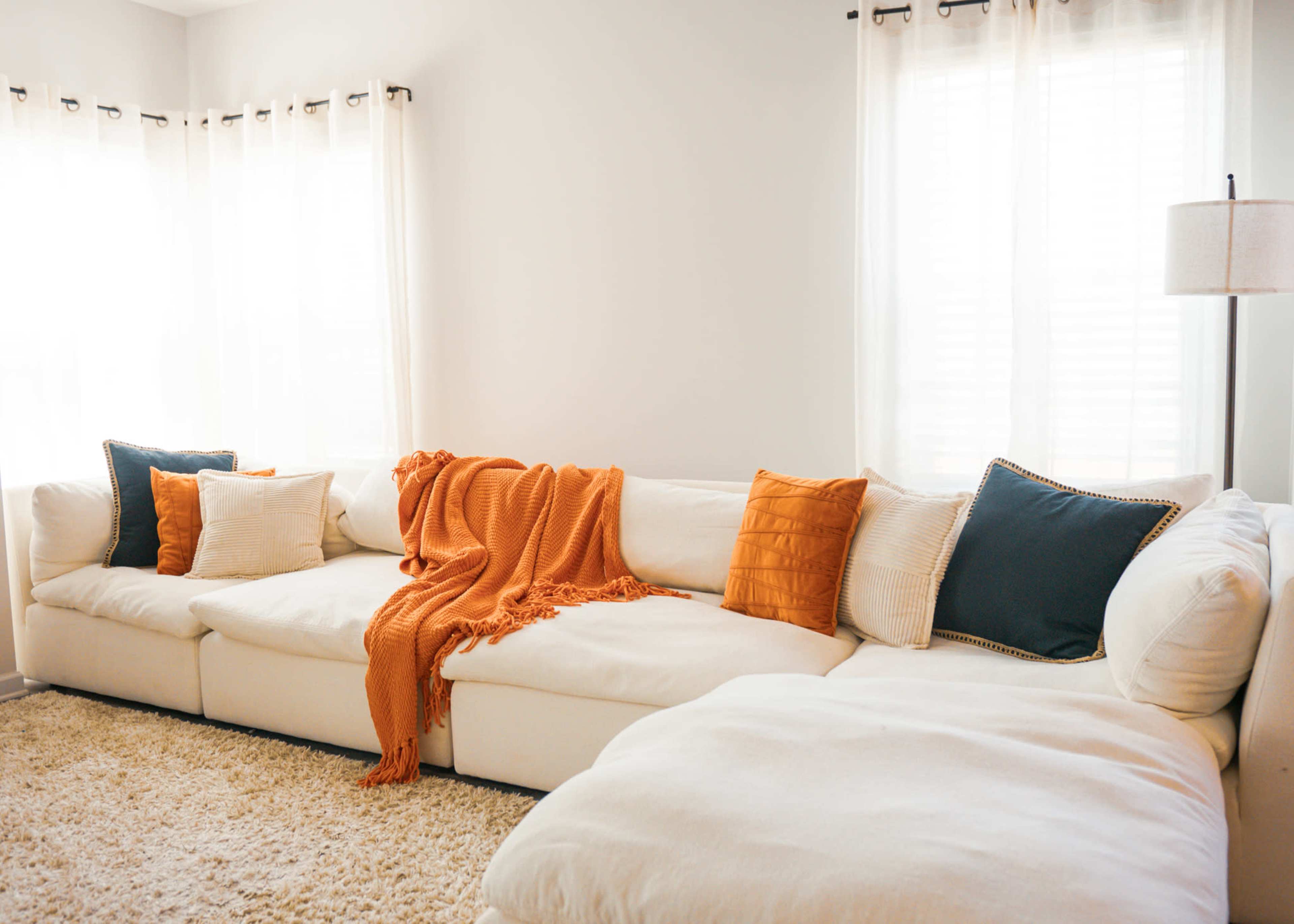 A white sectional sofa with orange and blue pillows is positioned in a room lit by natural light through sheer curtains.