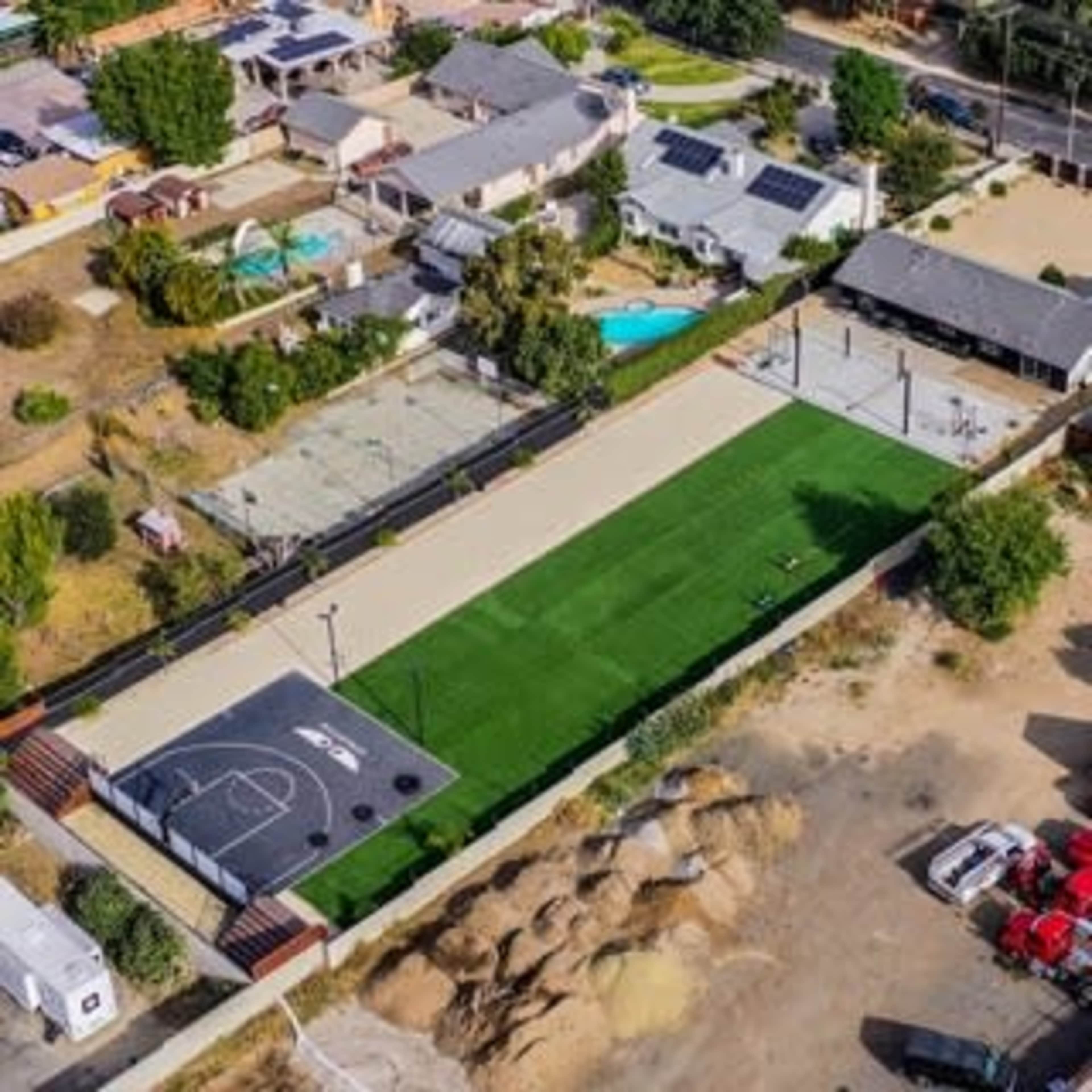 The image shows an aerial view of an outdoor recreational area featuring a basketball court, a turf field, and nearby residential properties.