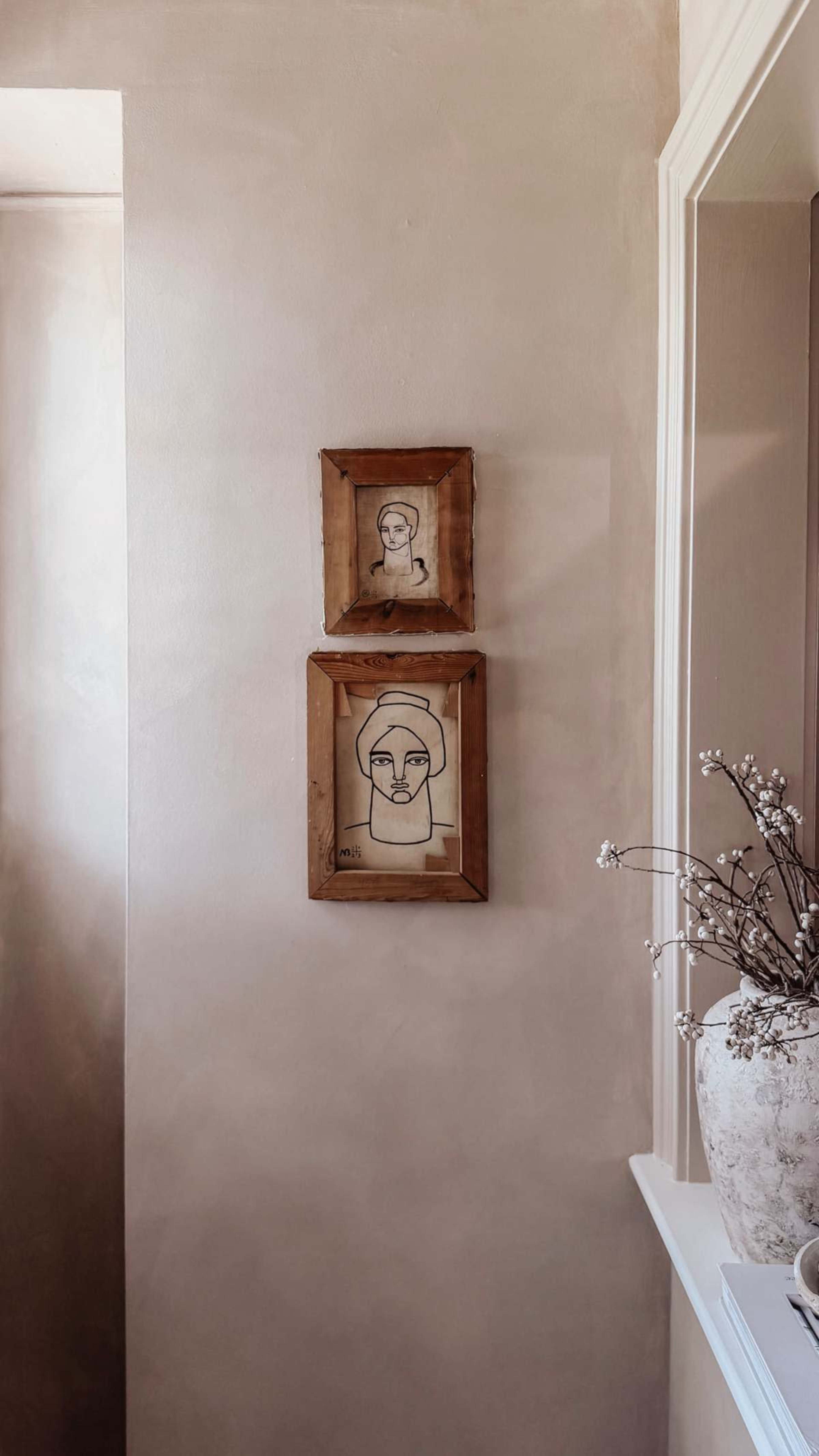 The image shows two framed drawings of faces hanging on a light-colored wall, with a vase of flowers on a ledge nearby.