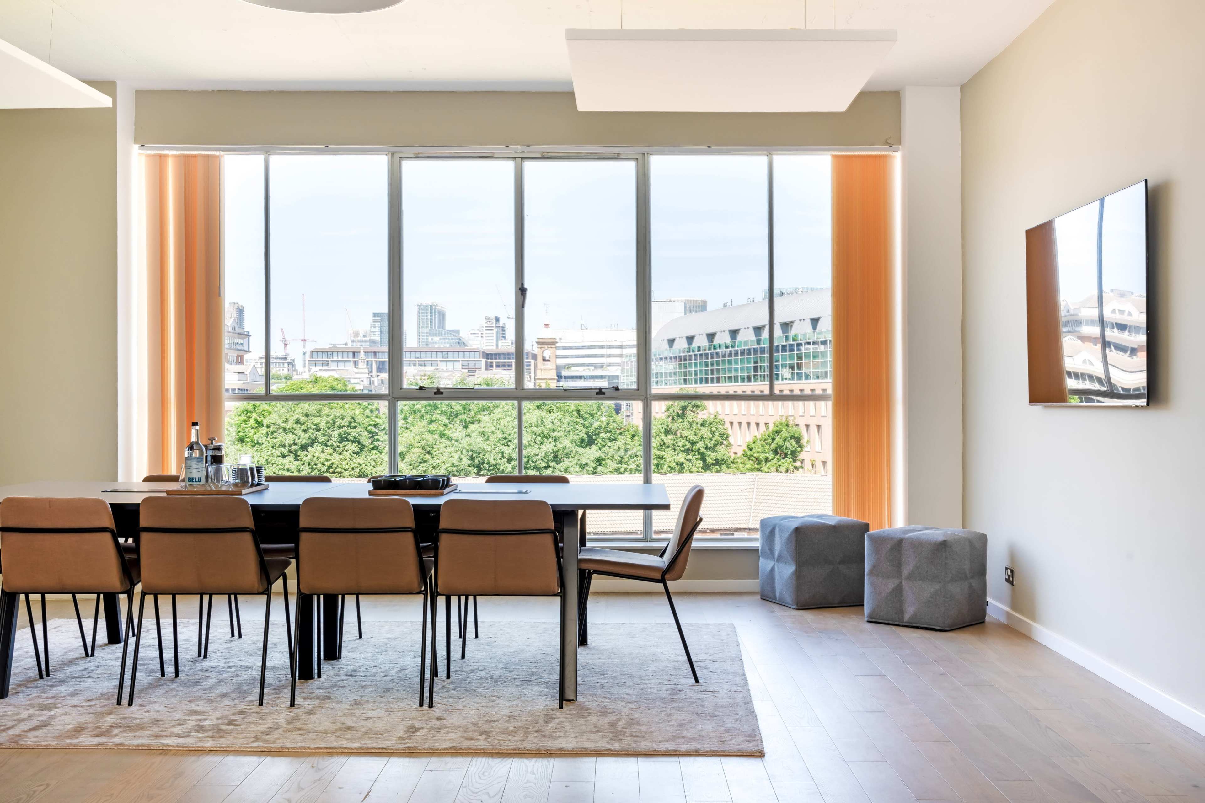 A modern dining area features a long table surrounded by chairs, with large windows offering a view of the city skyline.