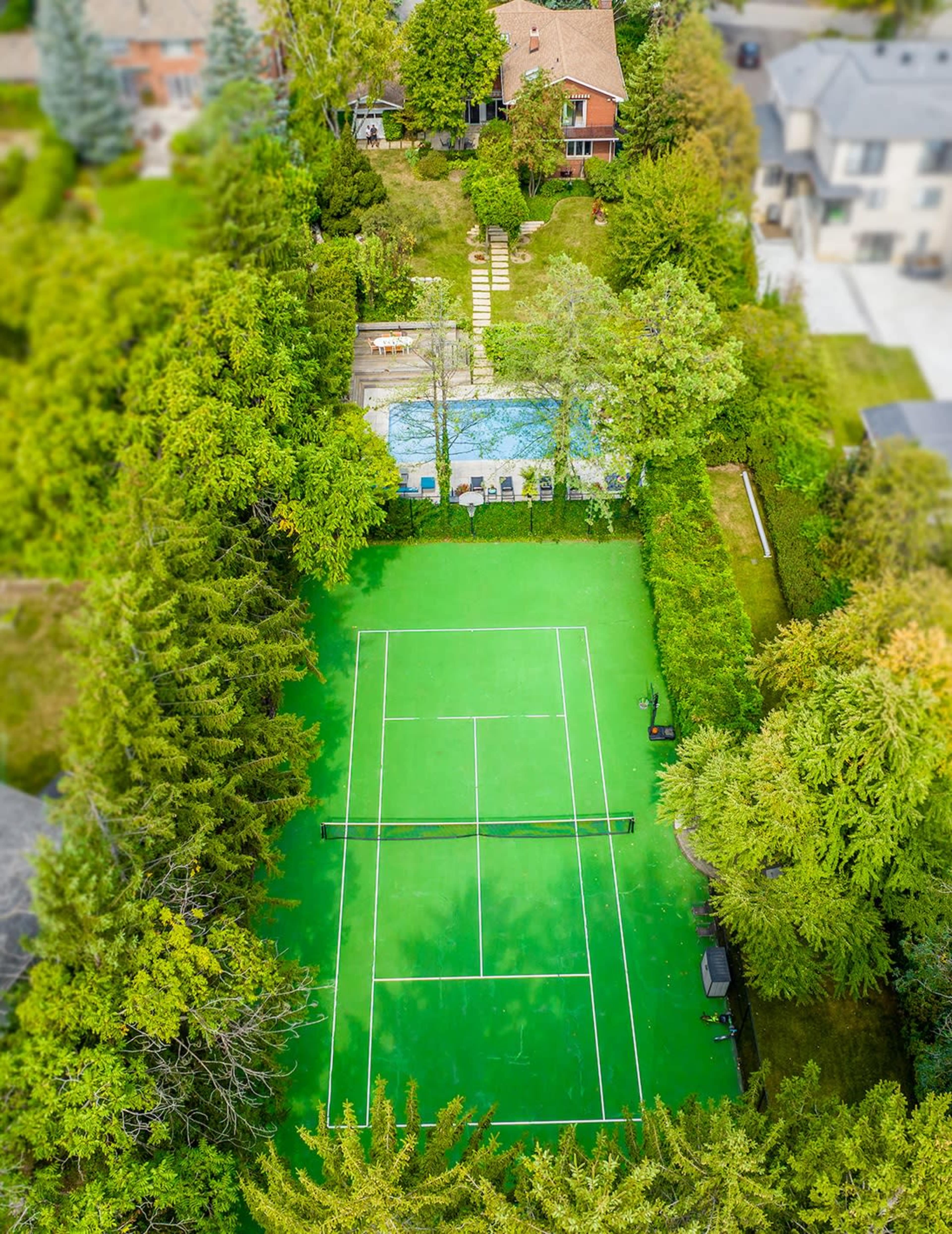 An aerial view of a home with a green tennis court surrounded by trees, a swimming pool, and a pathway leading to the house.