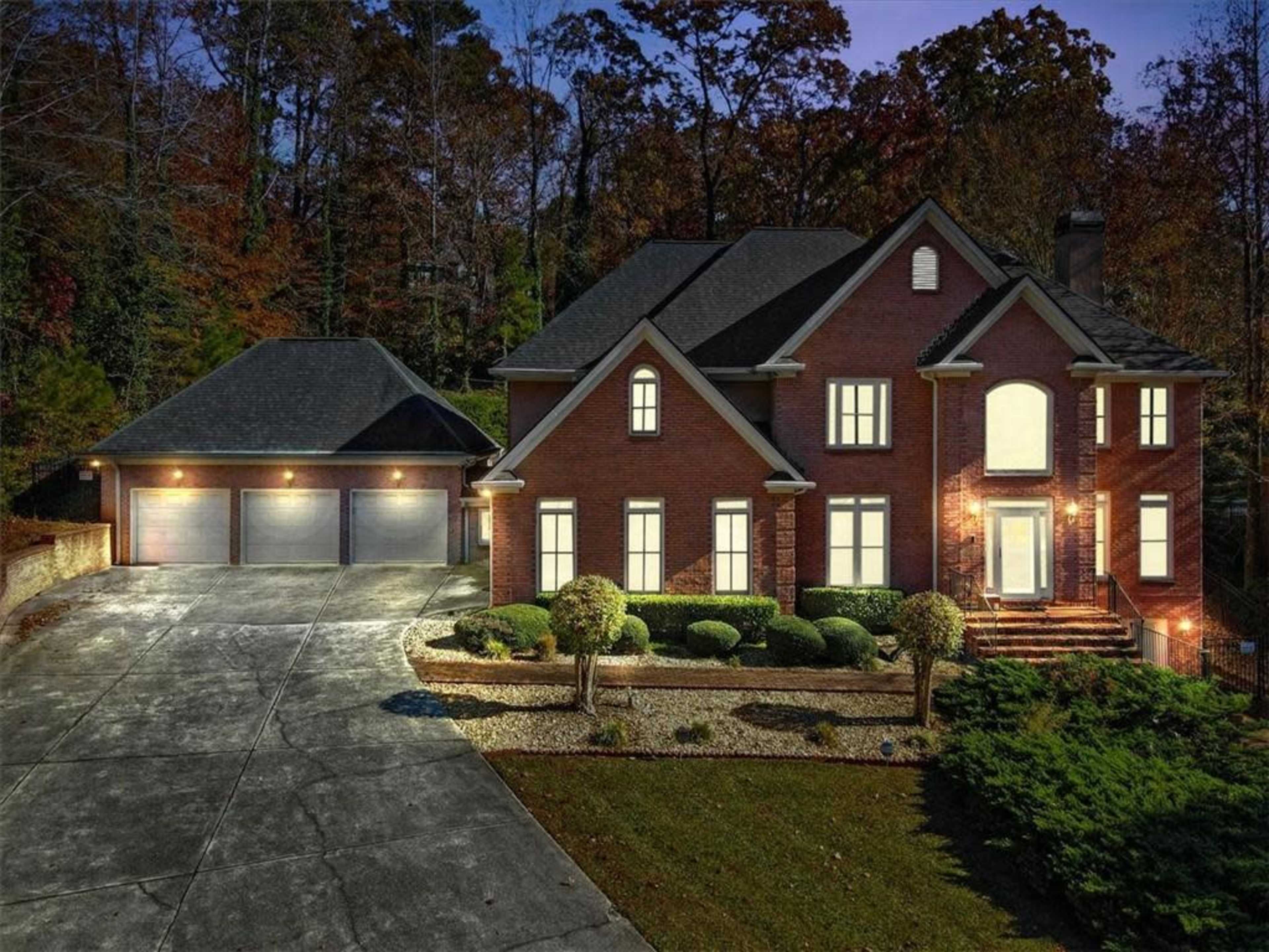 A large brick house sits on a landscaped lot with a three-car garage, illuminated by exterior lights against a backdrop of autumn trees.