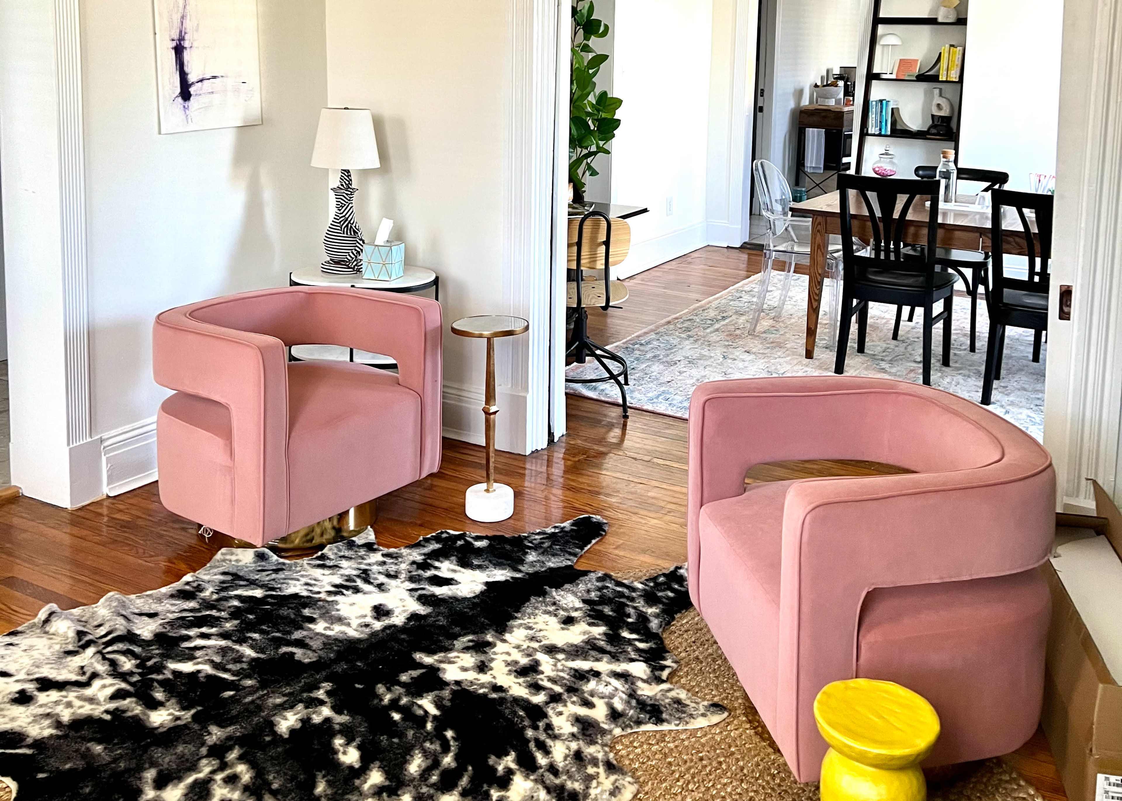 The image shows a bright living room with two pink armchairs, a small table, and a decorative black and white rug on the wooden floor.