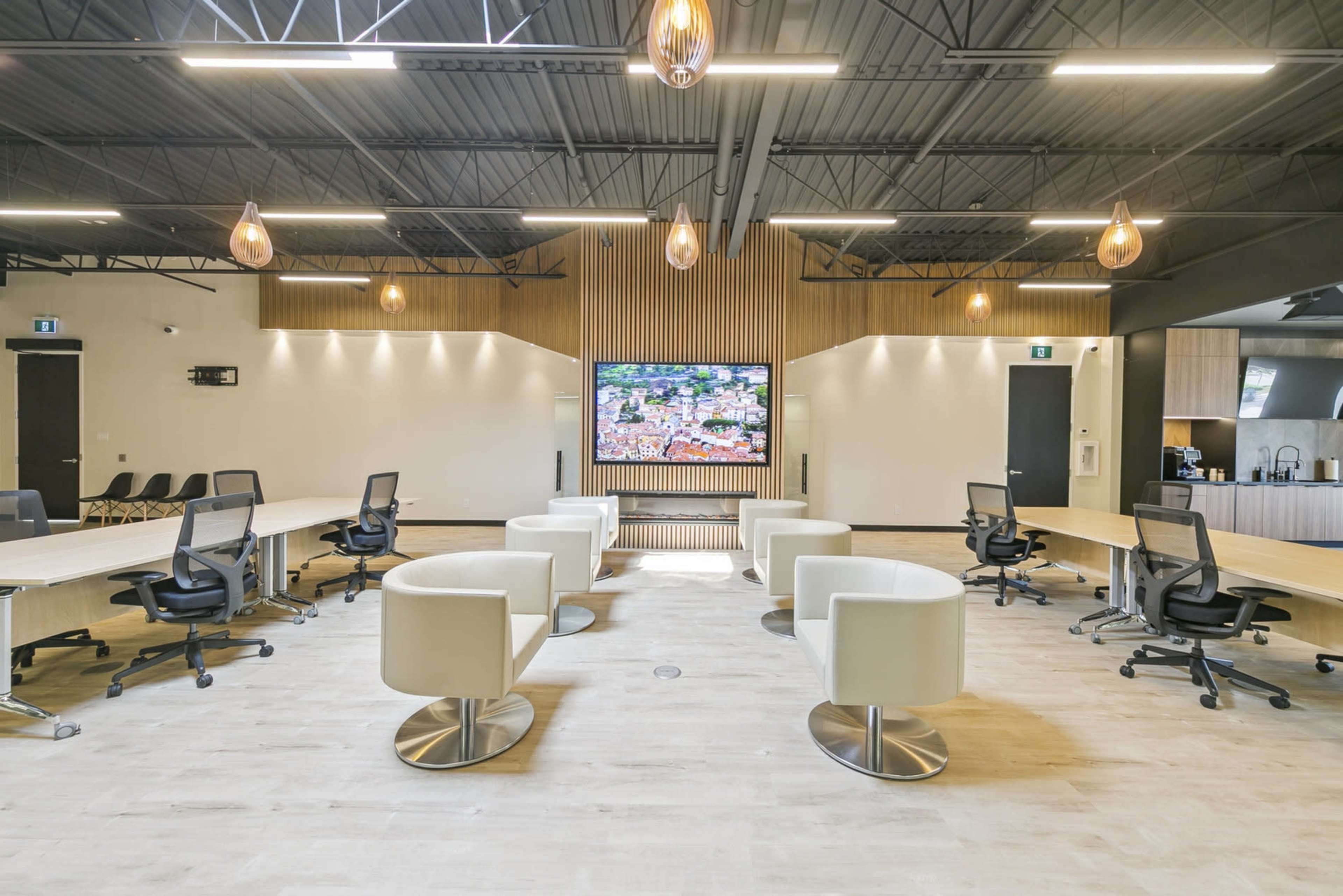 The image shows a modern, well-lit office space featuring circular seating arrangements around a central television, alongside long tables and office chairs.