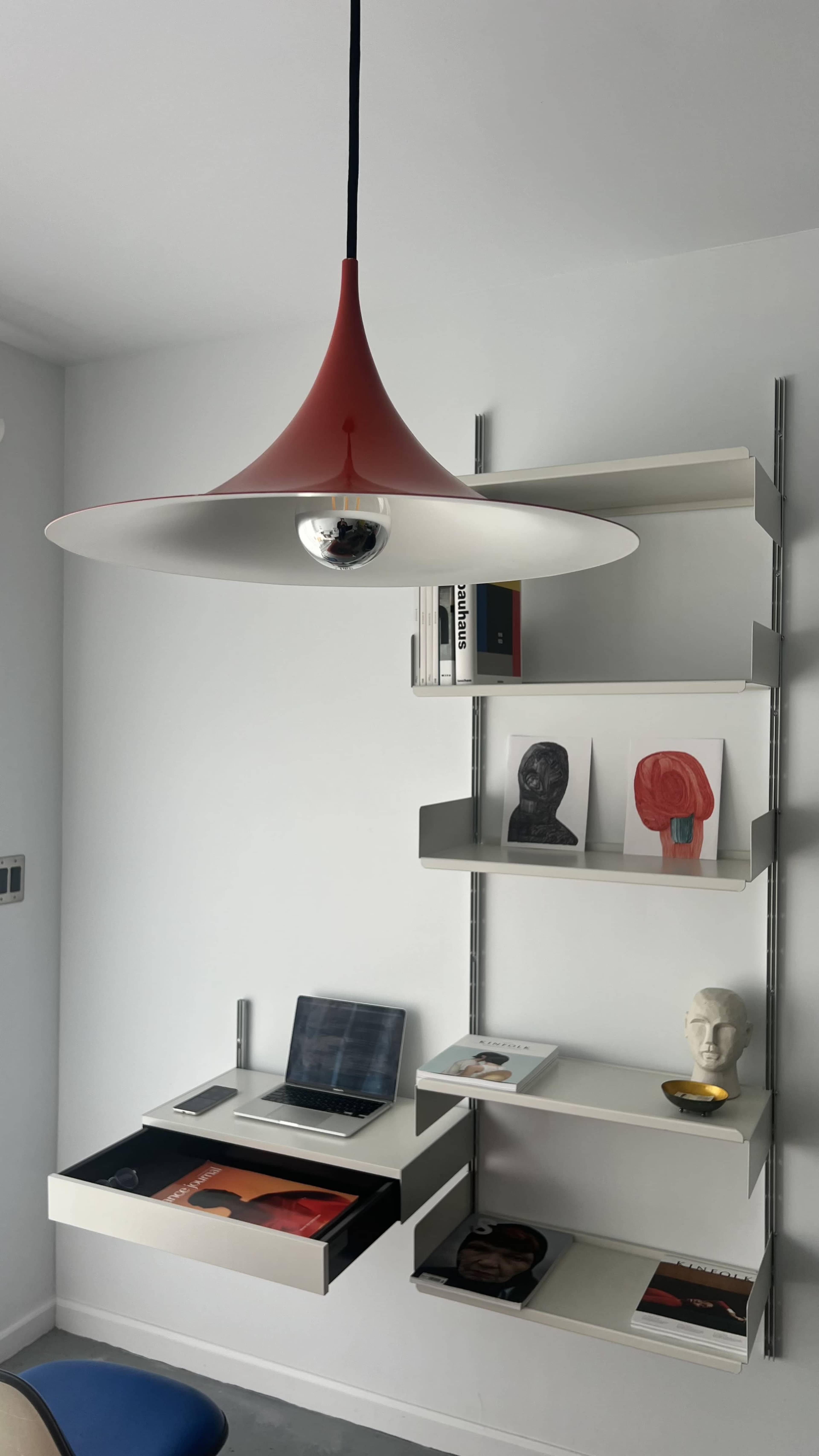 A red pendant lamp hangs above a minimalist desk featuring a laptop and two drawers, with a wall-mounted shelf displaying books and decorative artwork.