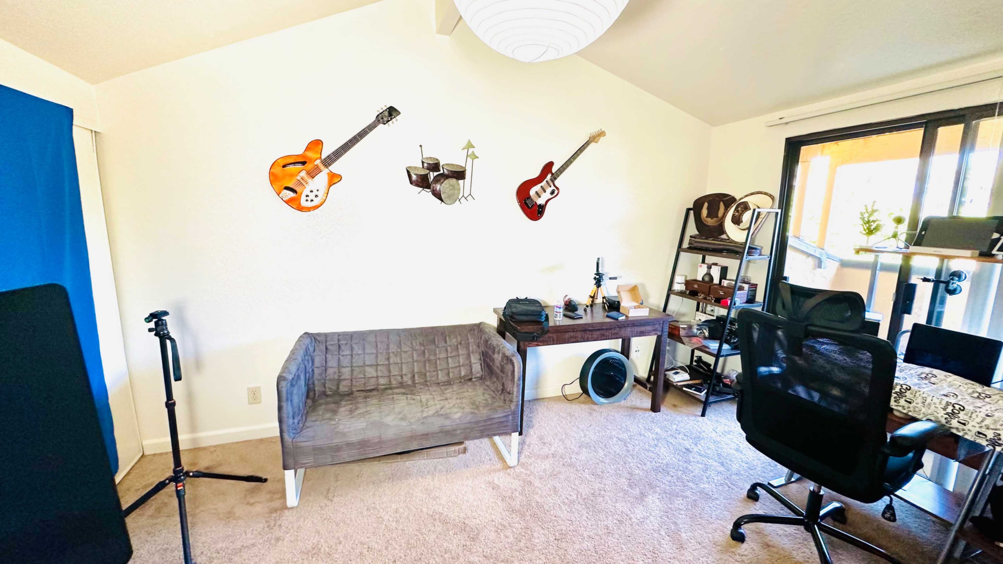 The room features a gray couch, a desk with various items, a black office chair, and three guitars mounted on the wall.