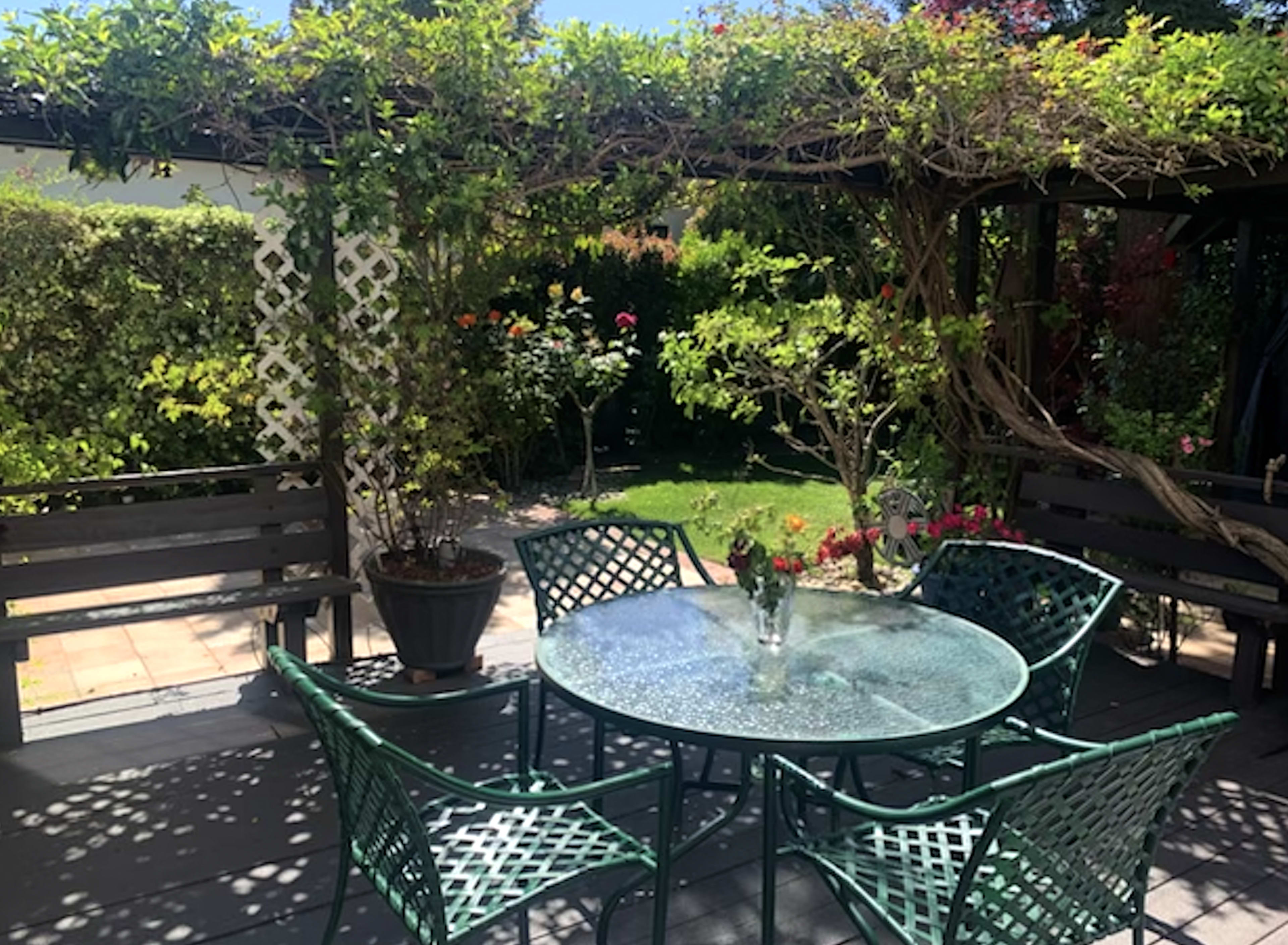 A round glass table surrounded by green chairs is set beneath a wooden pergola, with flowering plants and a lush garden in the background.