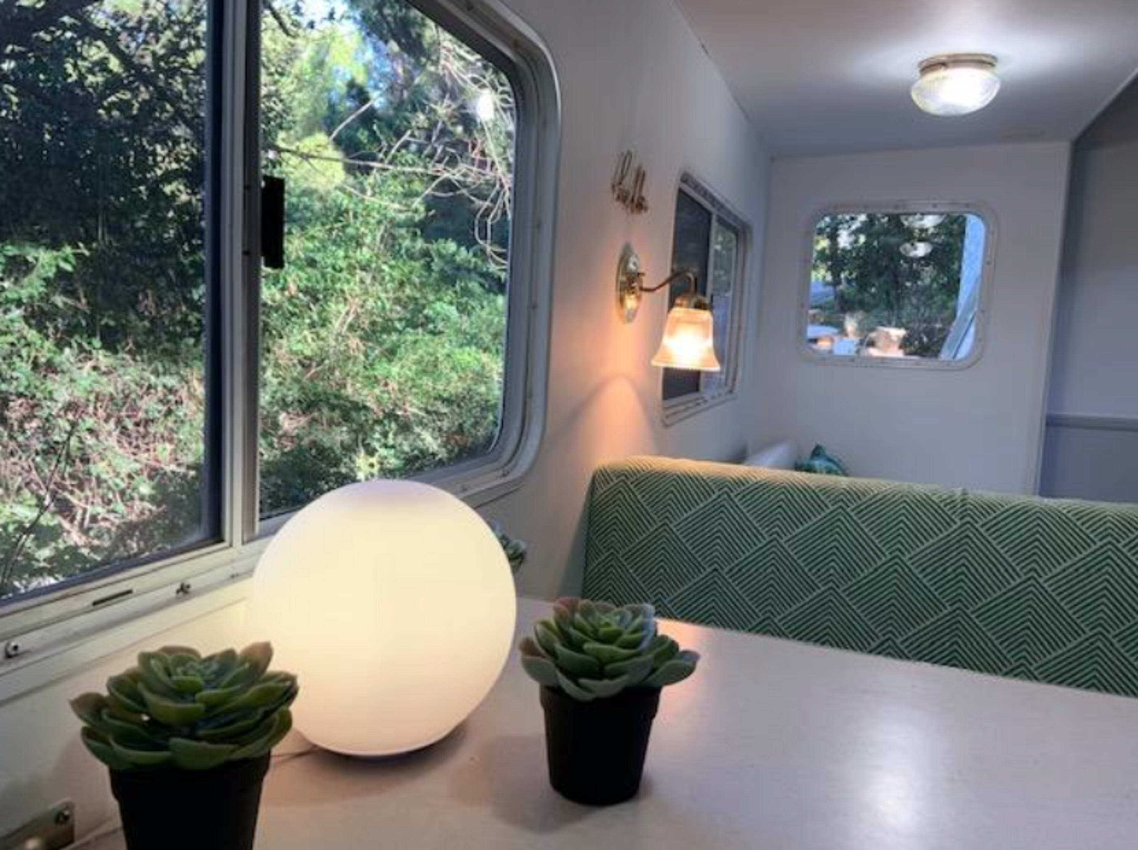 The interior of a small trailer features a round lamp, two succulent plants in pots, and a patterned green bench under a window overlooking greenery.