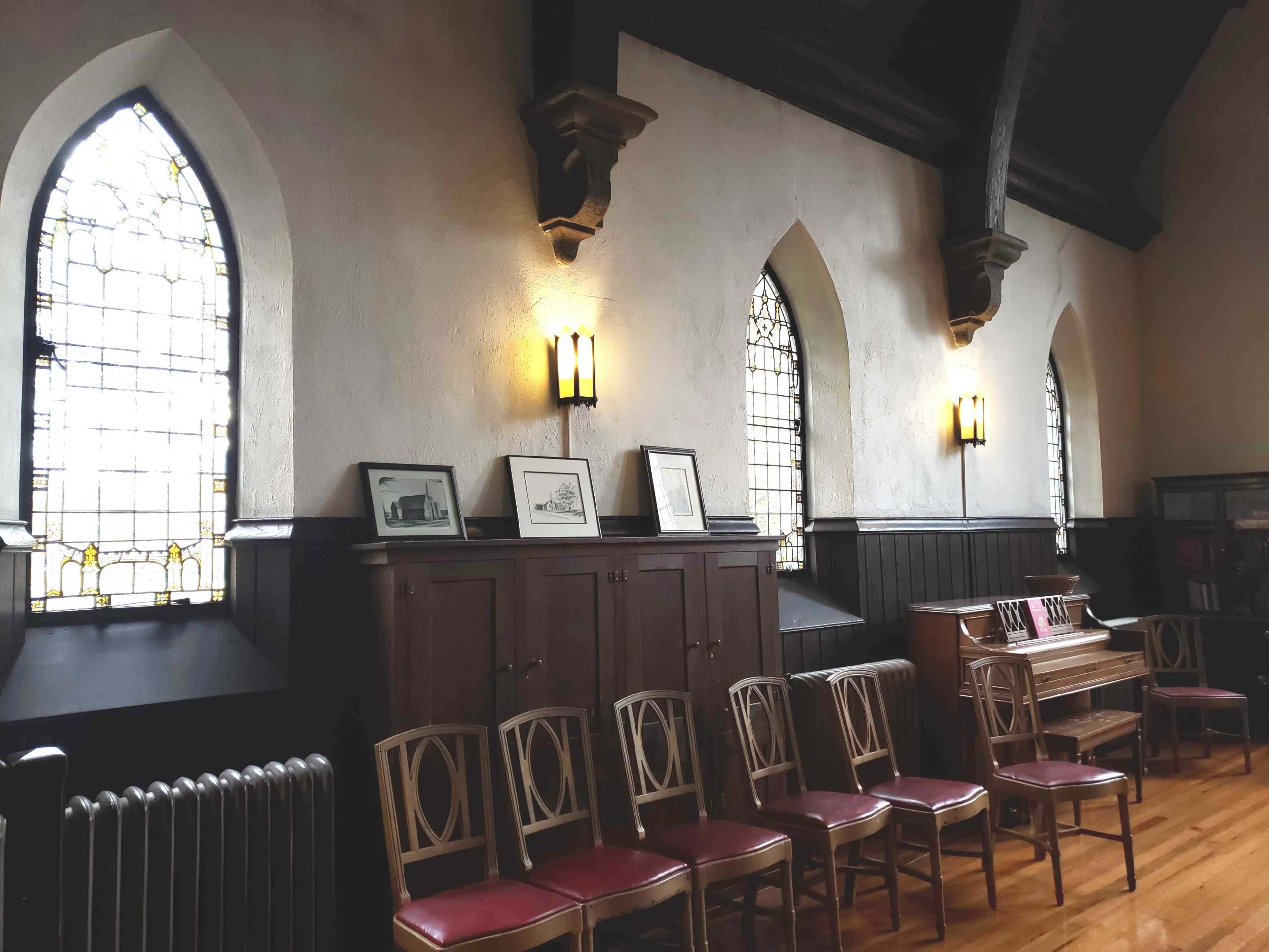 An interior of a room with stained glass windows, wooden paneling, a piano, and several chairs lined up against the wall.
