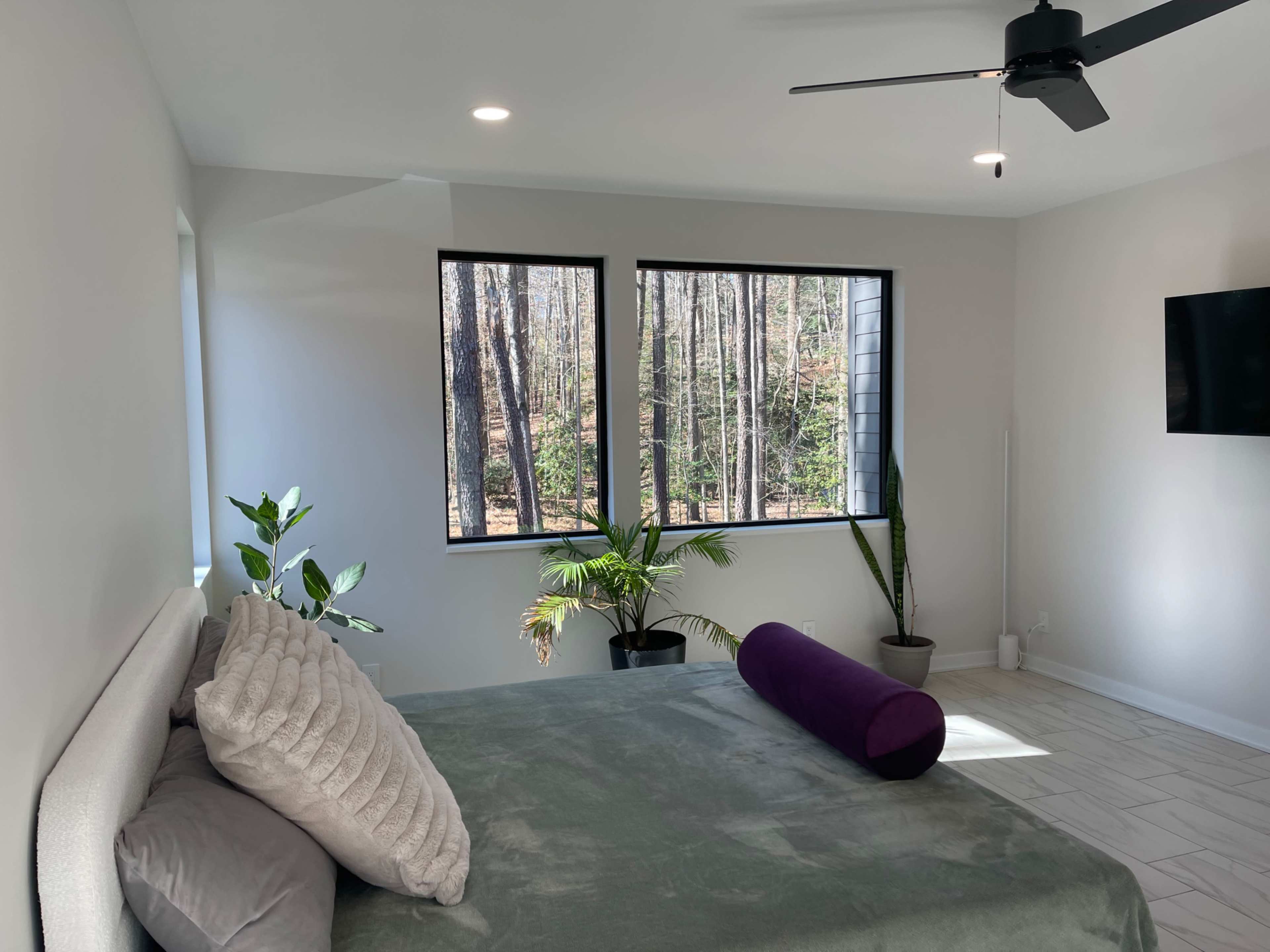 A modern bedroom features a bed with gray bedding and a purple bolster pillow, complemented by large windows that overlook a forested area.