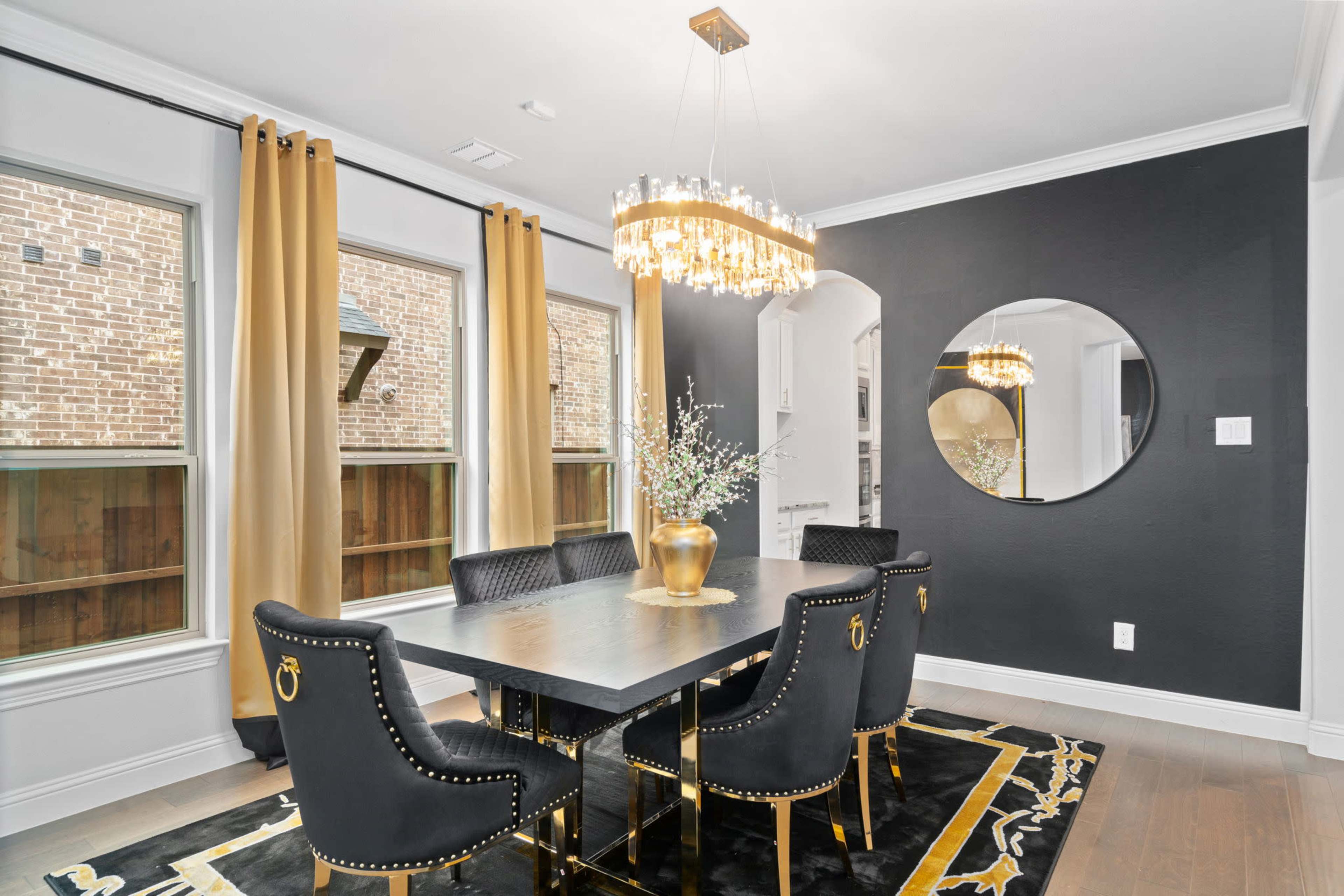 The dining room features a black table surrounded by six gold-accented chairs, a chandelier overhead, and large windows with golden curtains.