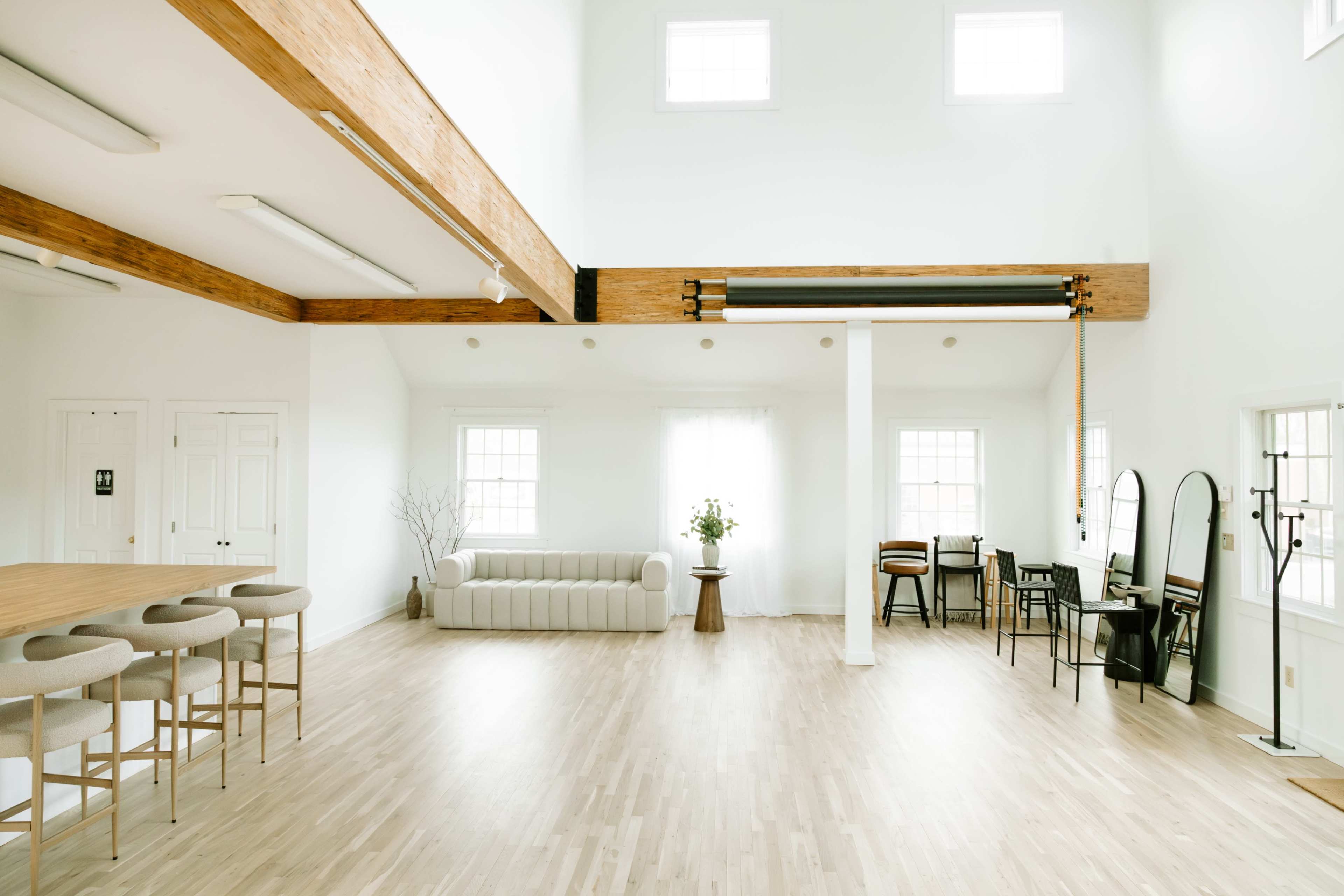 The image shows a spacious, modern interior with light wood flooring, a white sofa, a dining table with stools, and large windows.