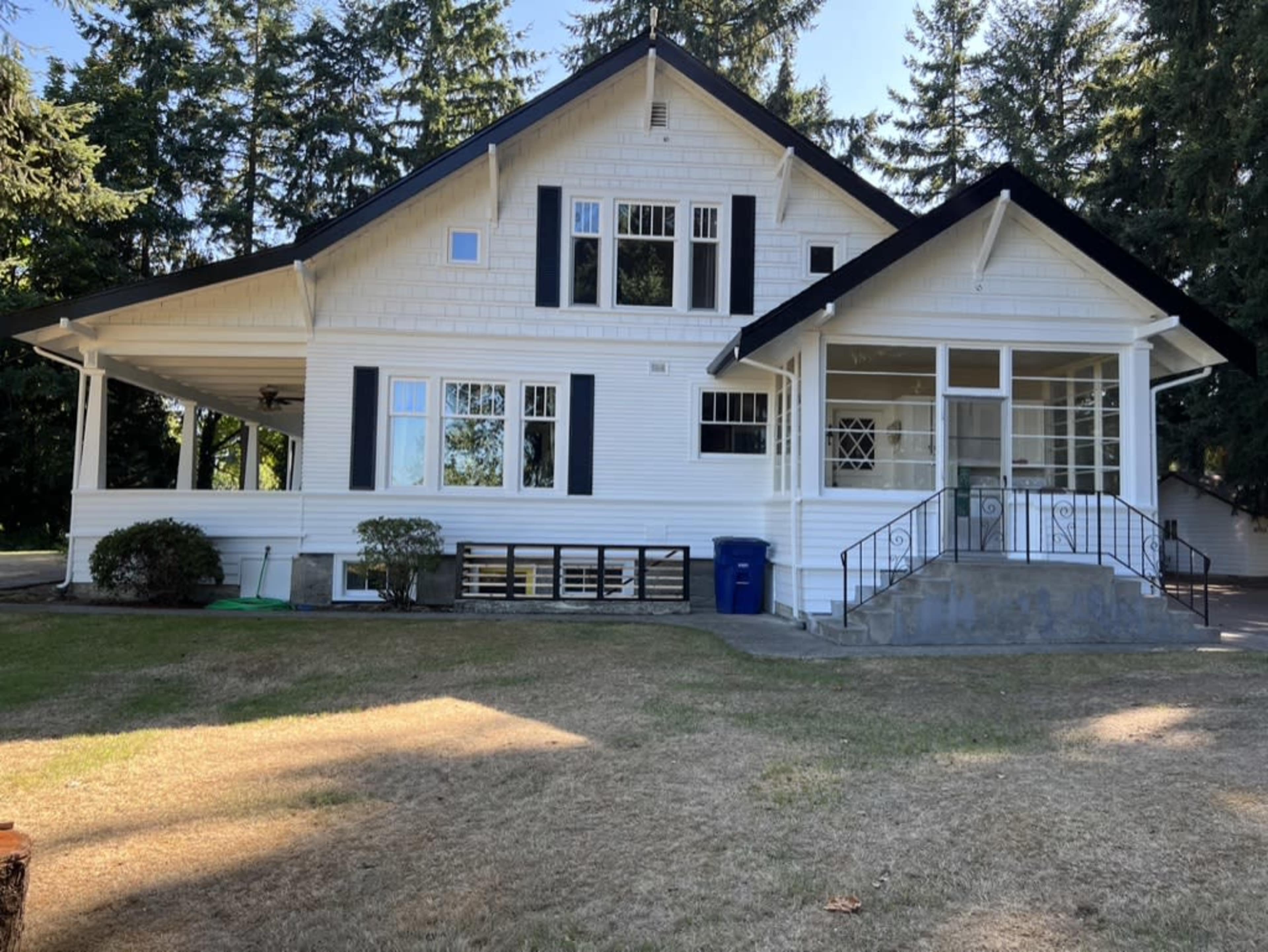 The image shows a large white house with a sloped roof, black shutters, and a porch, surrounded by trees and a lawn.
