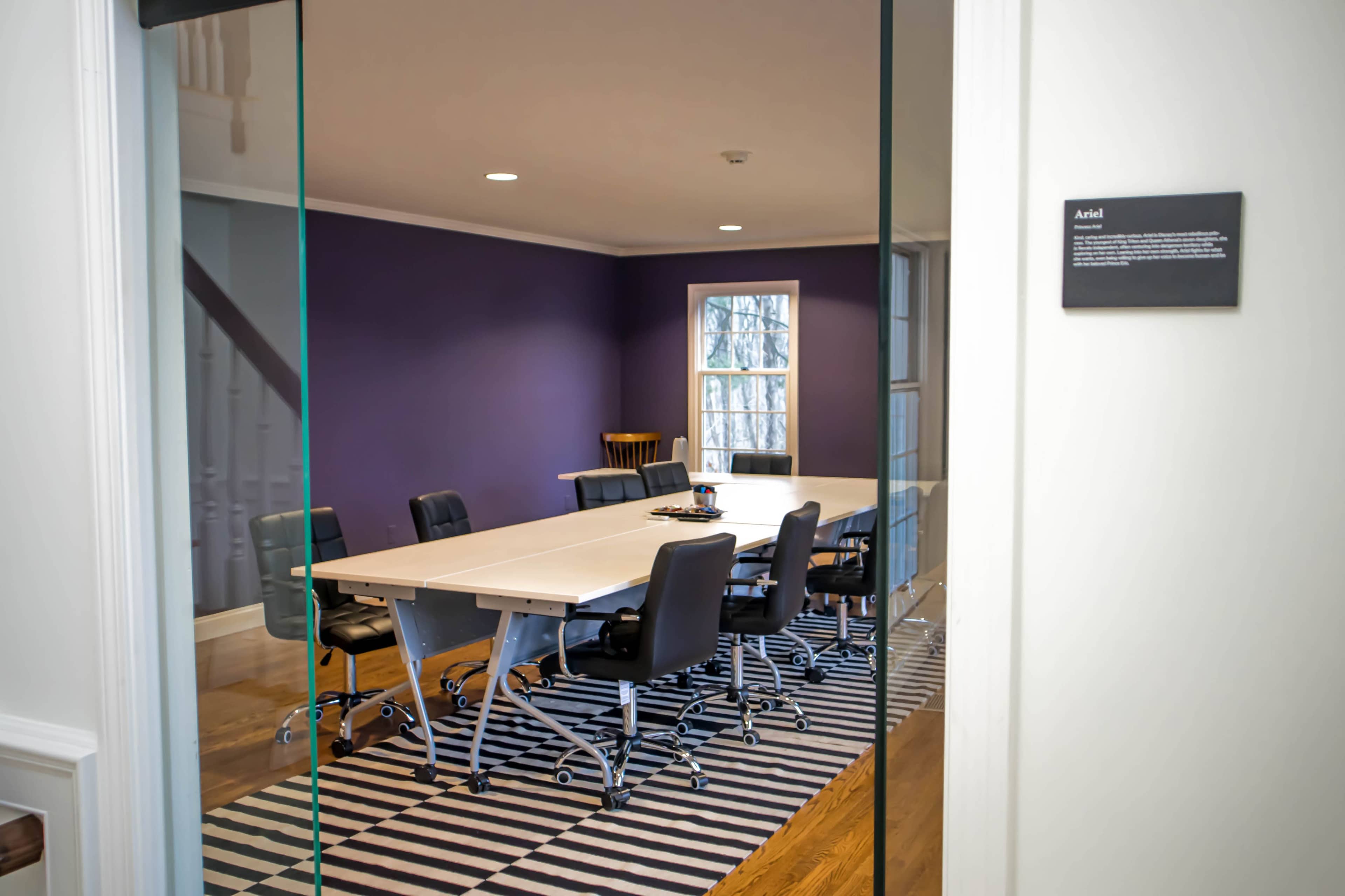 A modern, minimalist workspace with a table, chairs, and a purple wall in the background.