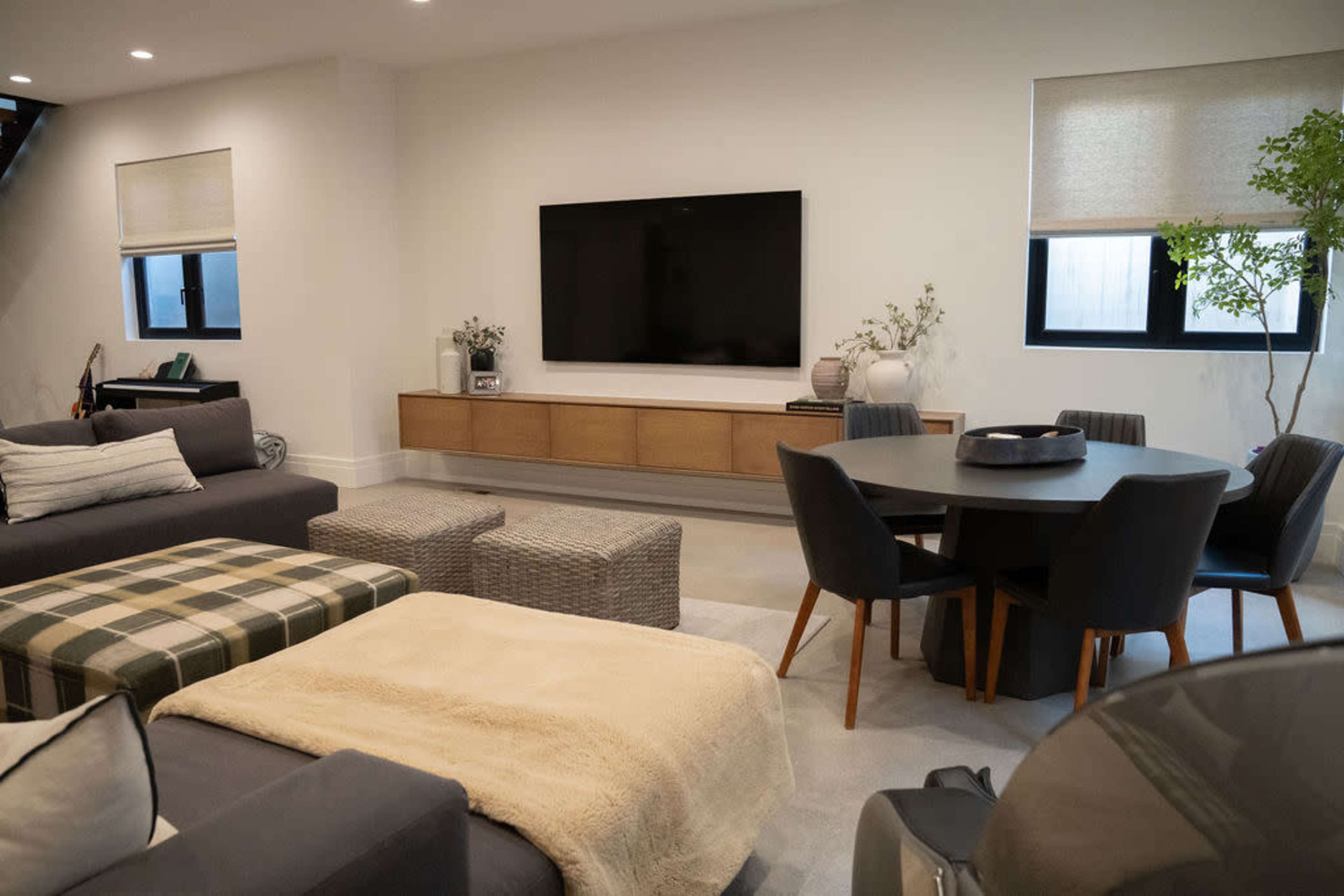 A modern living room features a large television mounted on a light-colored wall, with a mix of seating arrangements, including a sofa and a round dining table, surrounded by neutral tones and natural materials.