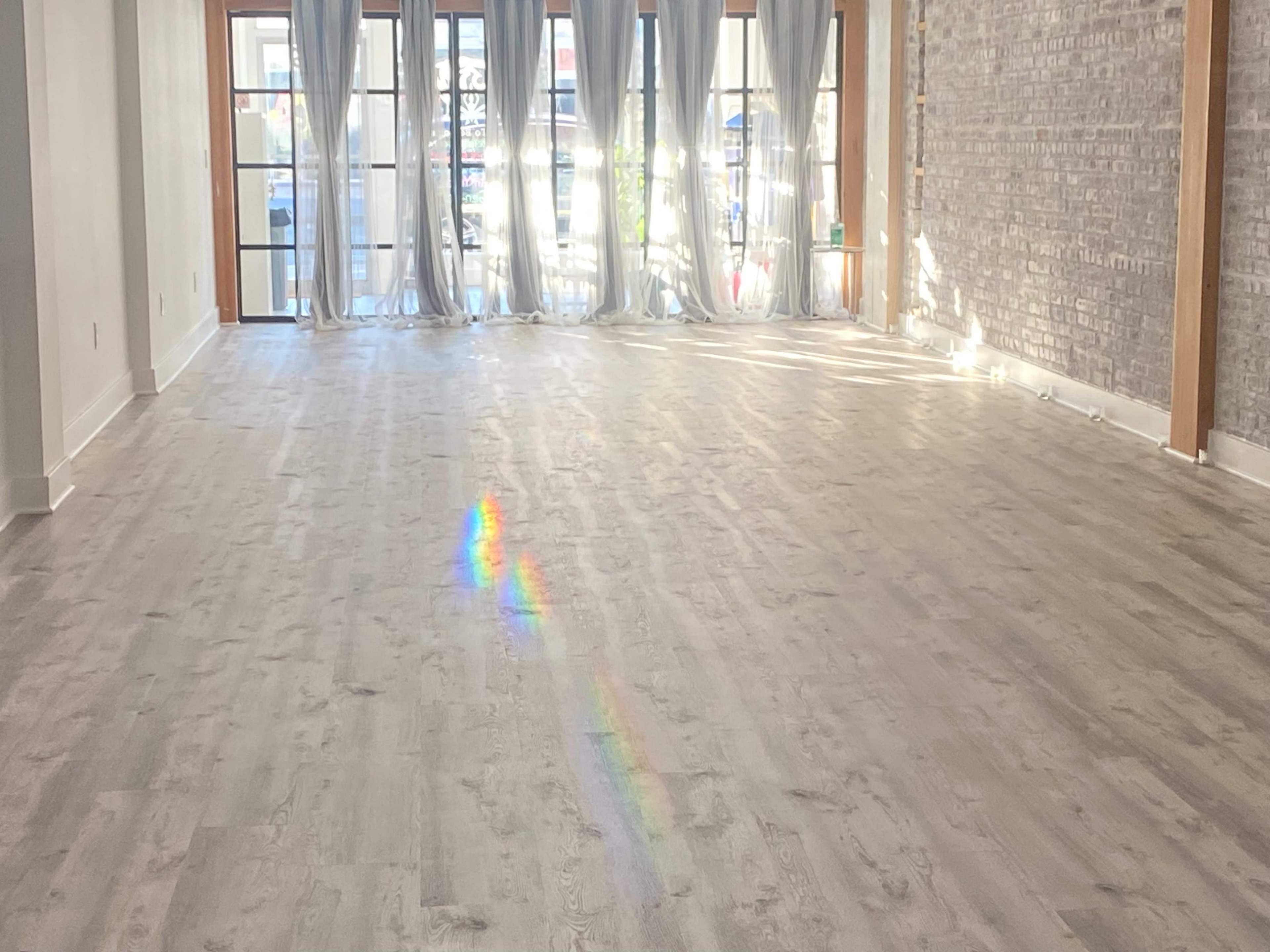 A spacious room with light-colored wooden flooring and large windows covered by sheer curtains, allowing natural light to enter.