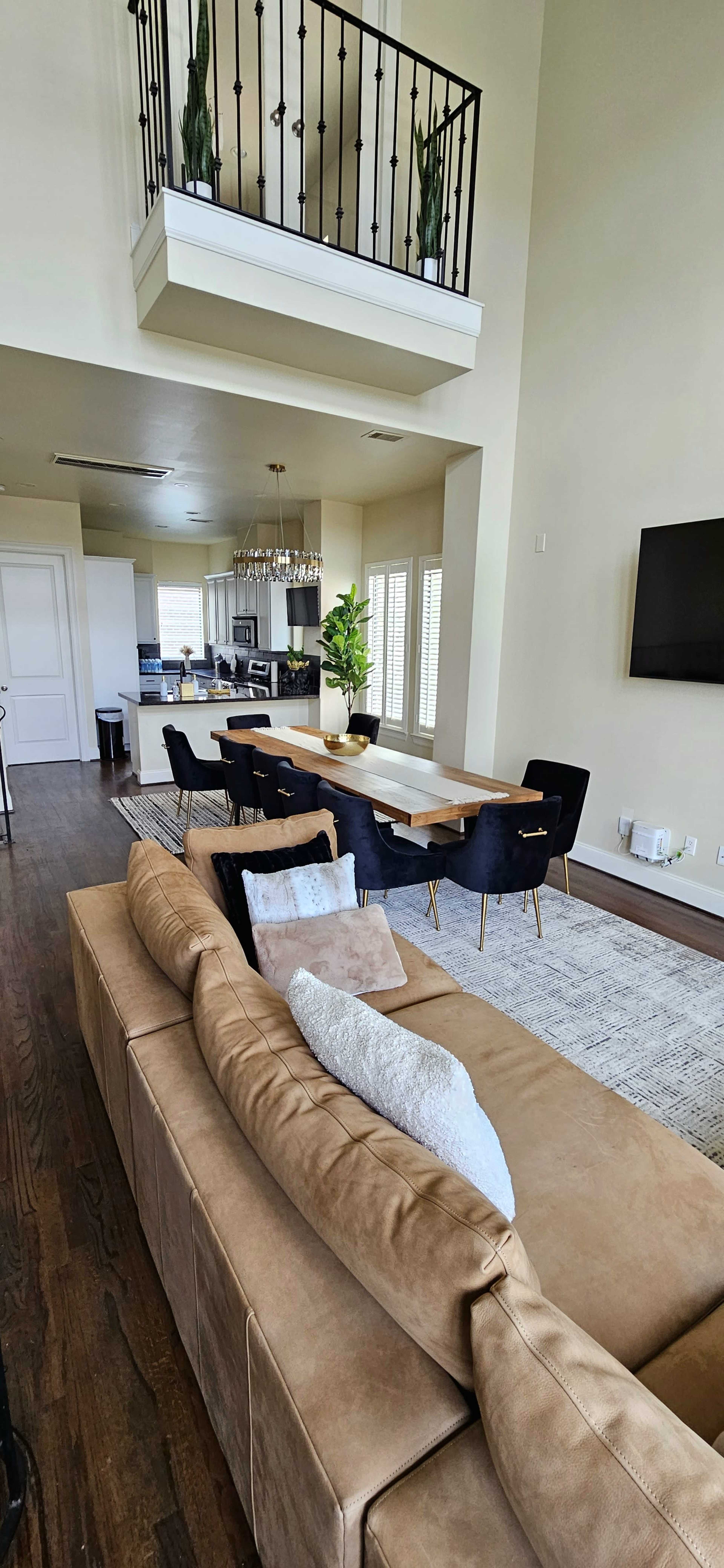 A spacious living area with a tan sectional sofa, a dining table surrounded by black chairs, a modern kitchen in the background, and large windows letting in light.