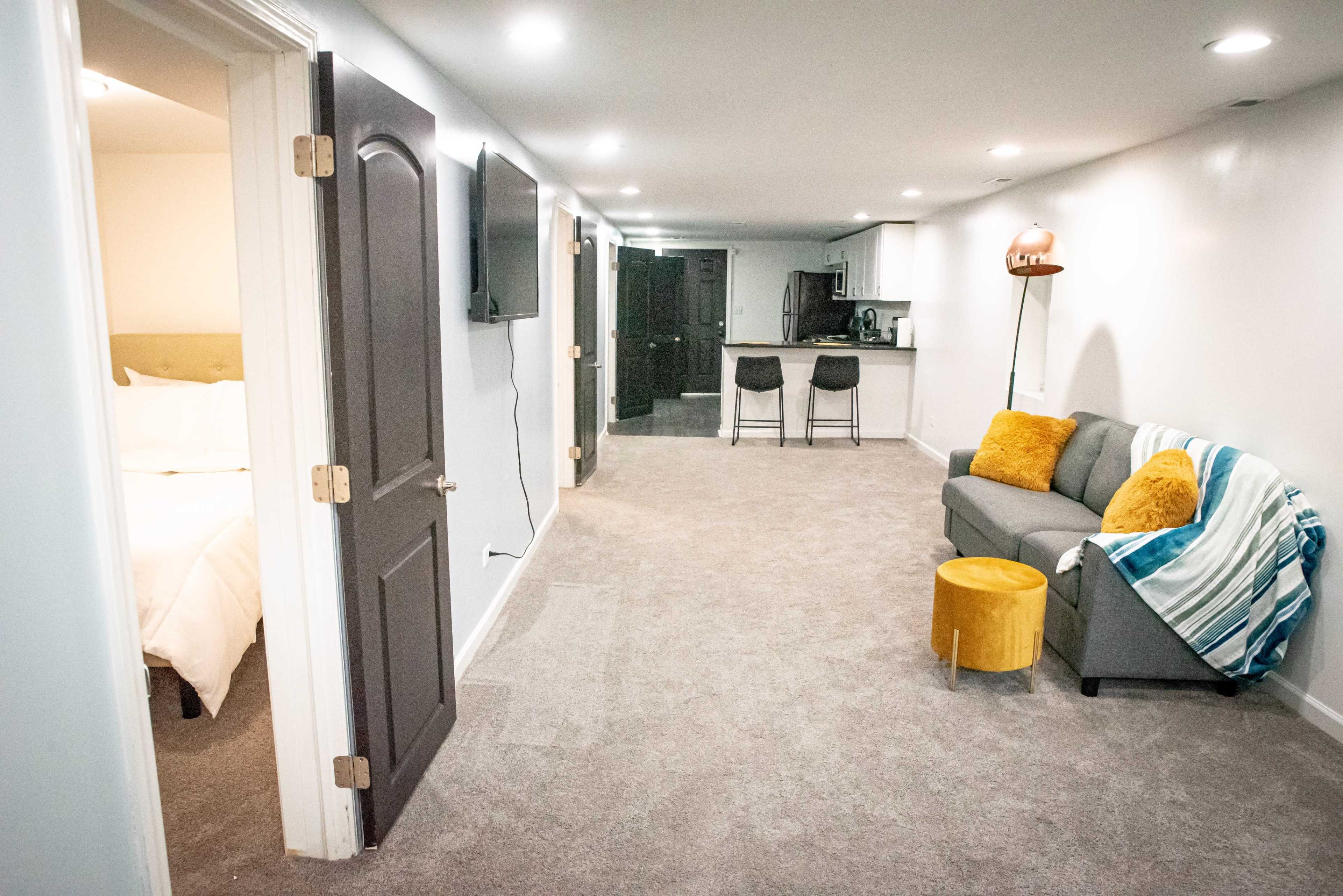 A well-lit, carpeted hallway leading to a kitchen area, with a door opening to a bedroom on the left and a sofa with an accent chair on the right.