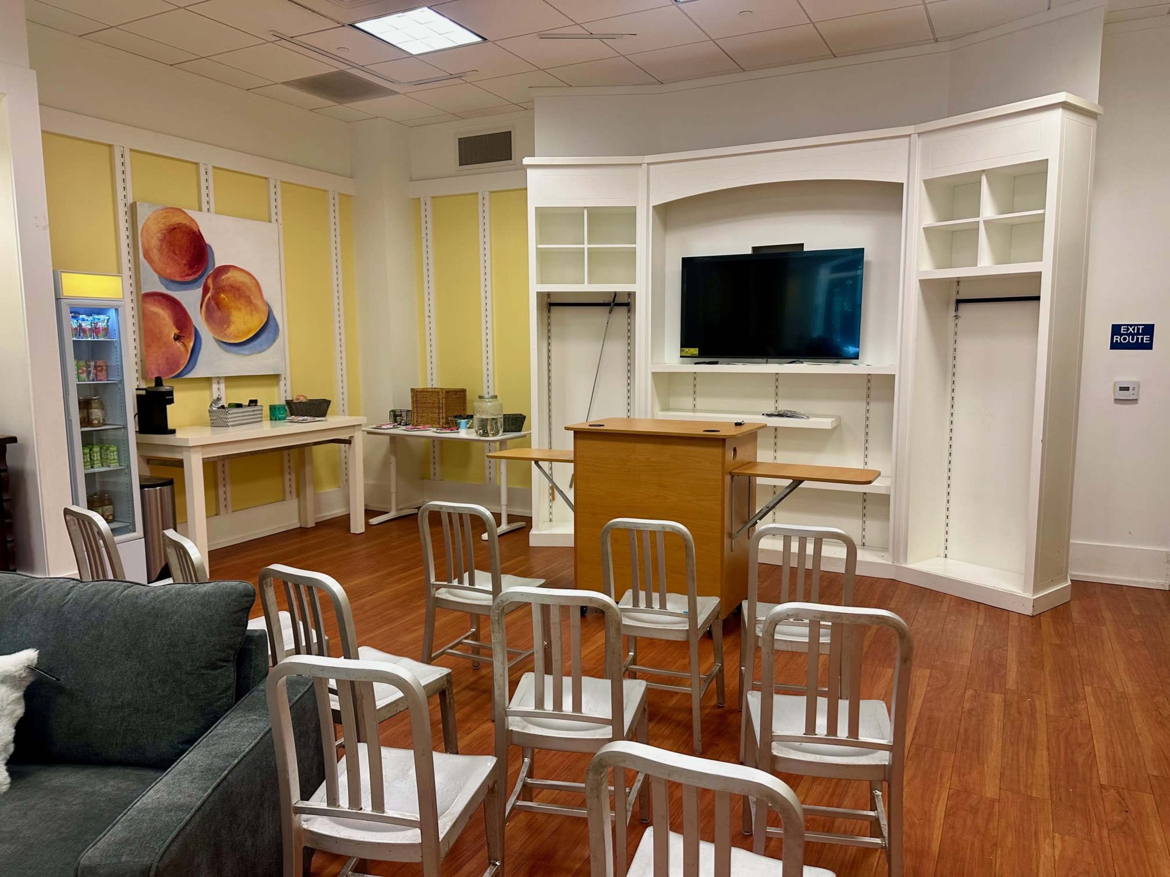 The image shows a room set up with several metal chairs facing a wooden table in front of a television, with a few shelves and a fruit painting on the wall.