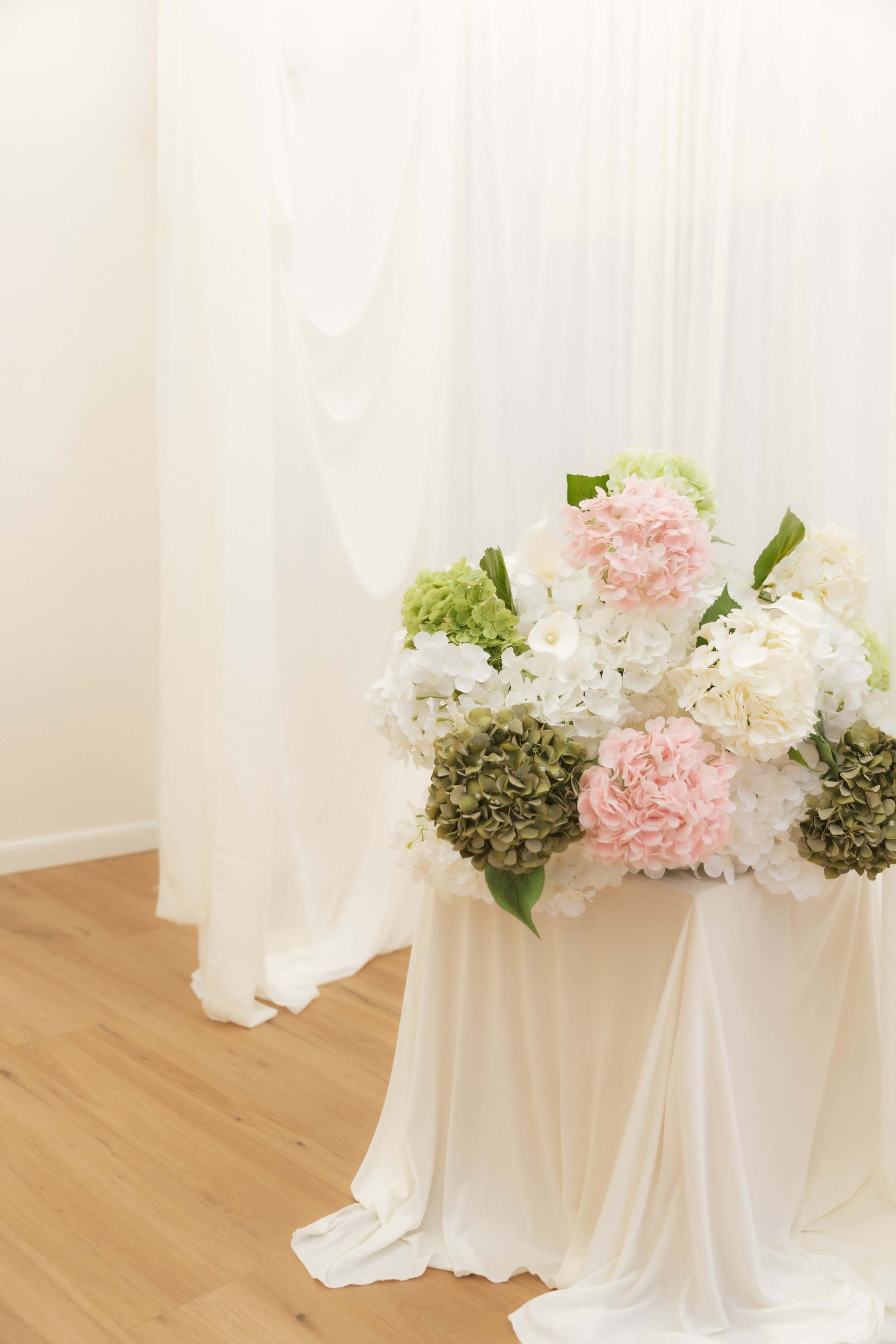 A floral arrangement featuring pink, white, and green flowers is displayed in front of white drapery and wooden flooring.