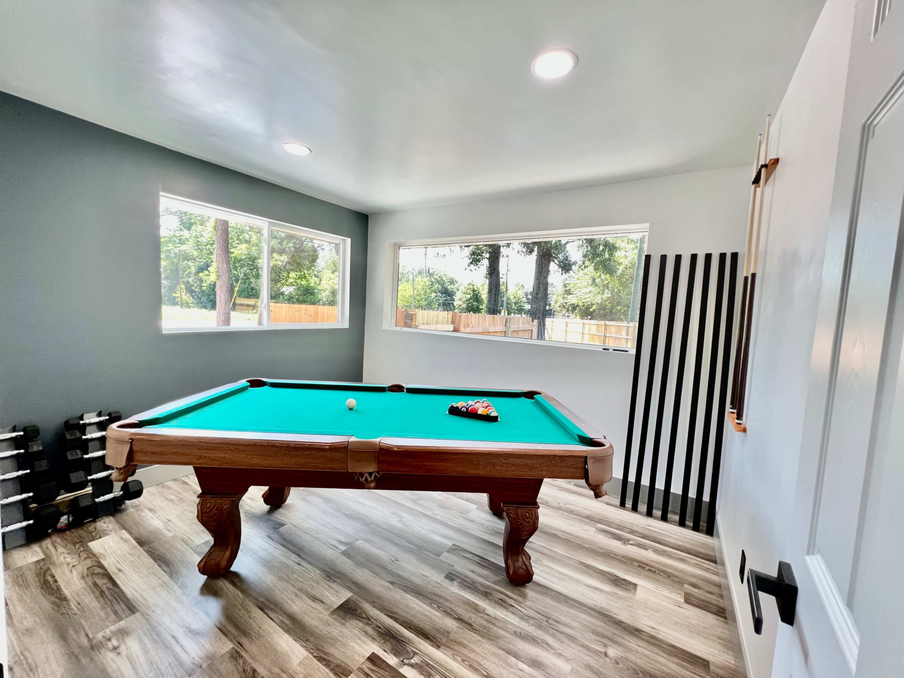 A bright game room featuring a pool table, surrounded by windows and a set of dumbbells in the corner.