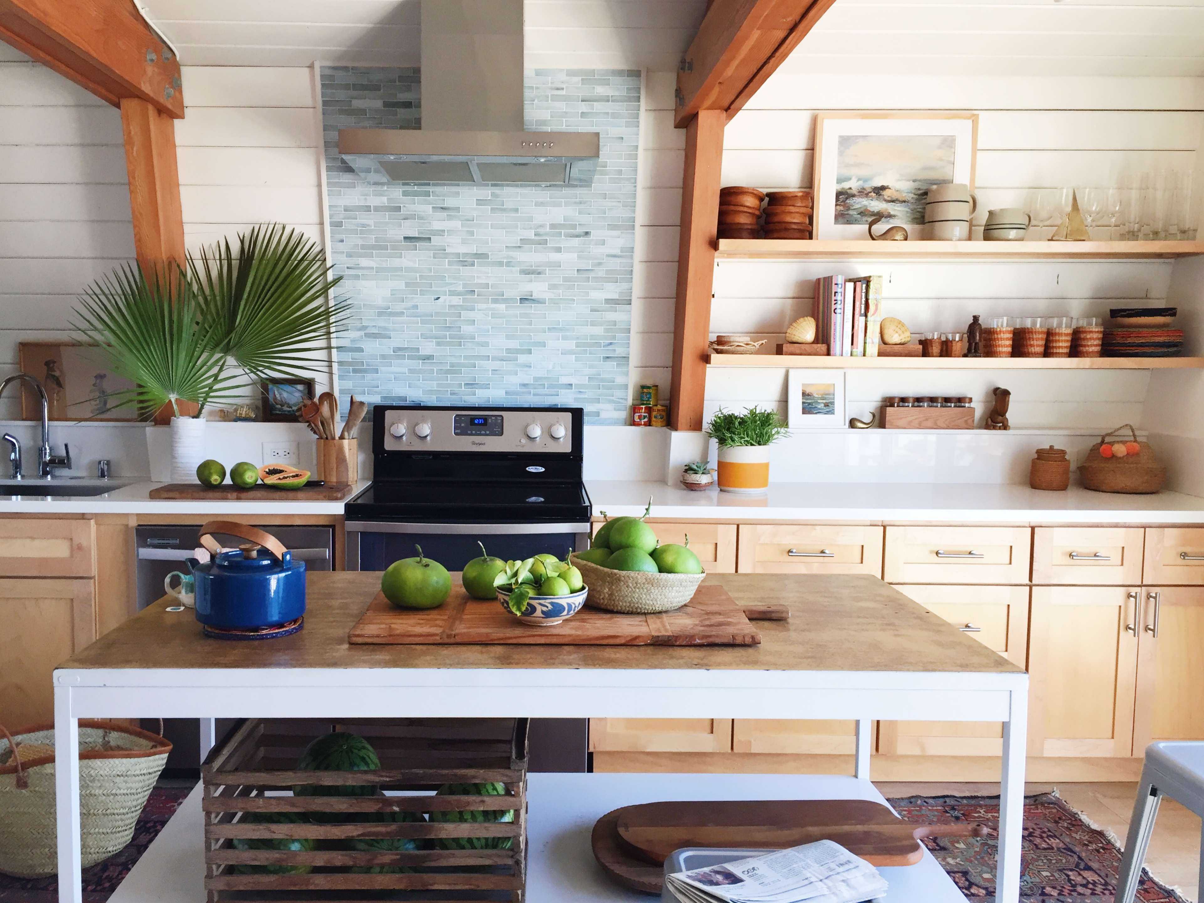 A modern kitchen features a central island with green apples, a stove, wooden shelves, and decorative plants in a bright, airy space.