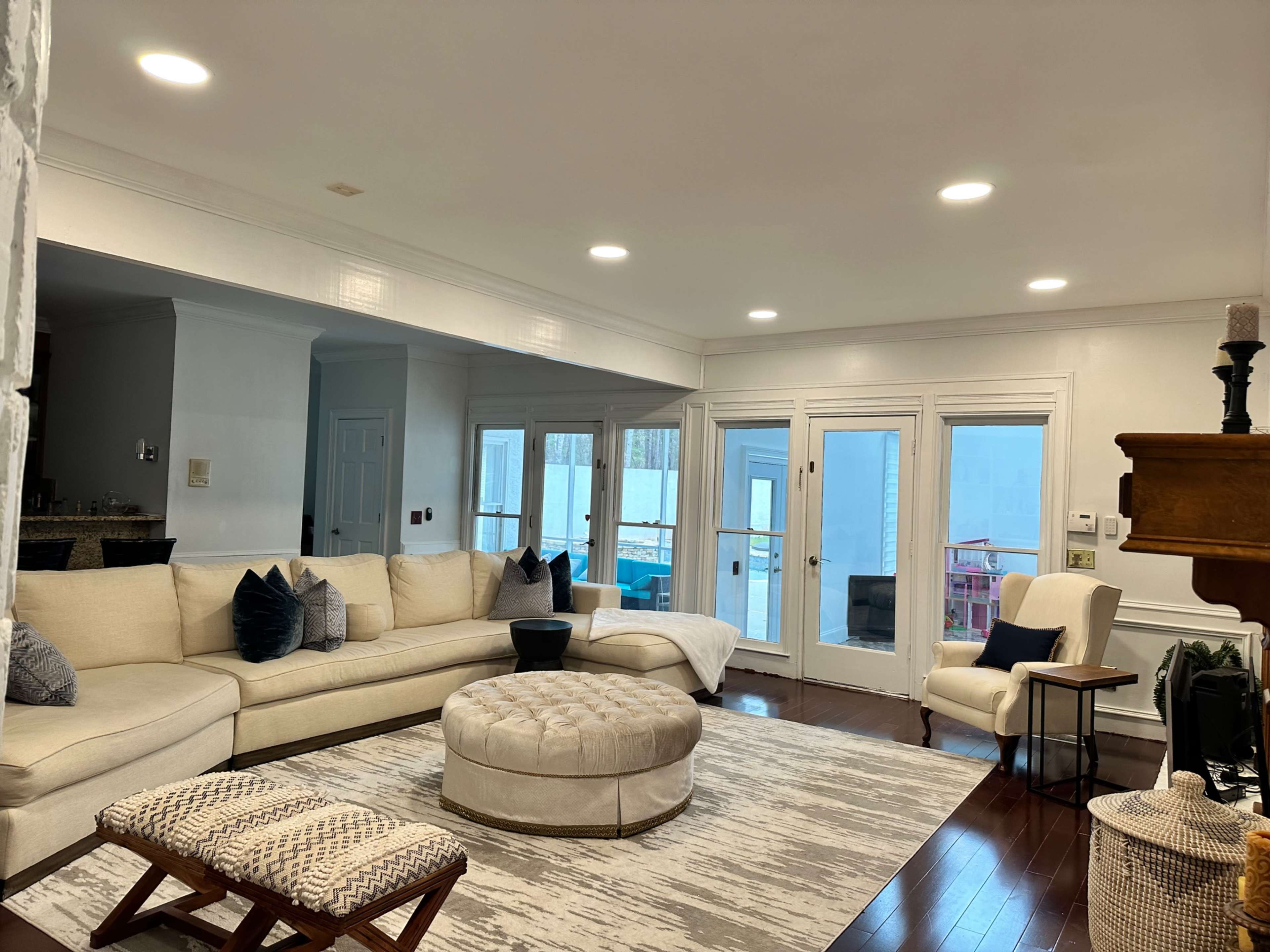The image shows a spacious living room with a large sectional sofa, an ottoman in the center, and glass doors leading to an outdoor area.