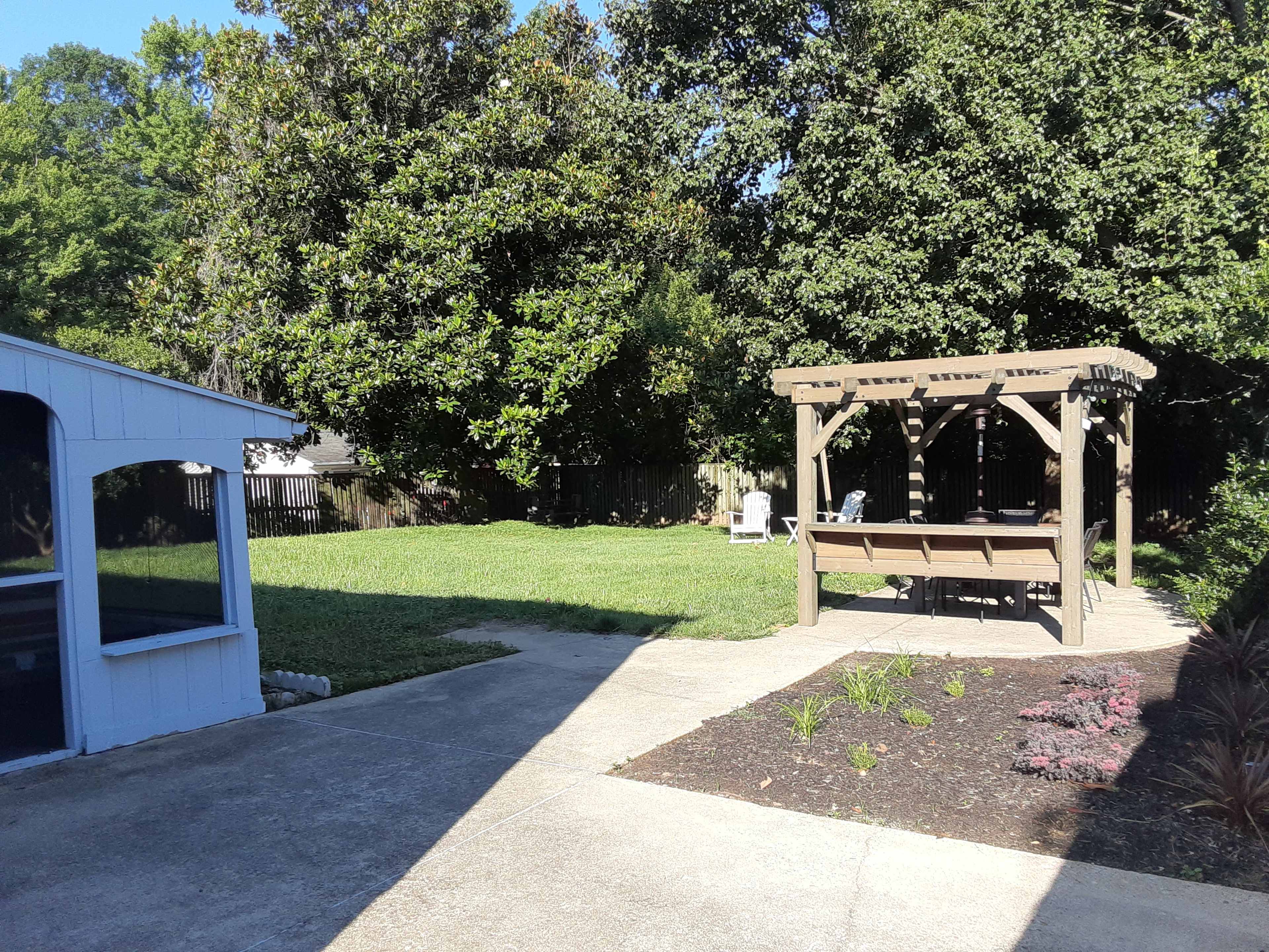 A grassy backyard with a wooden pergola and a patio area, bordered by trees and flower beds.