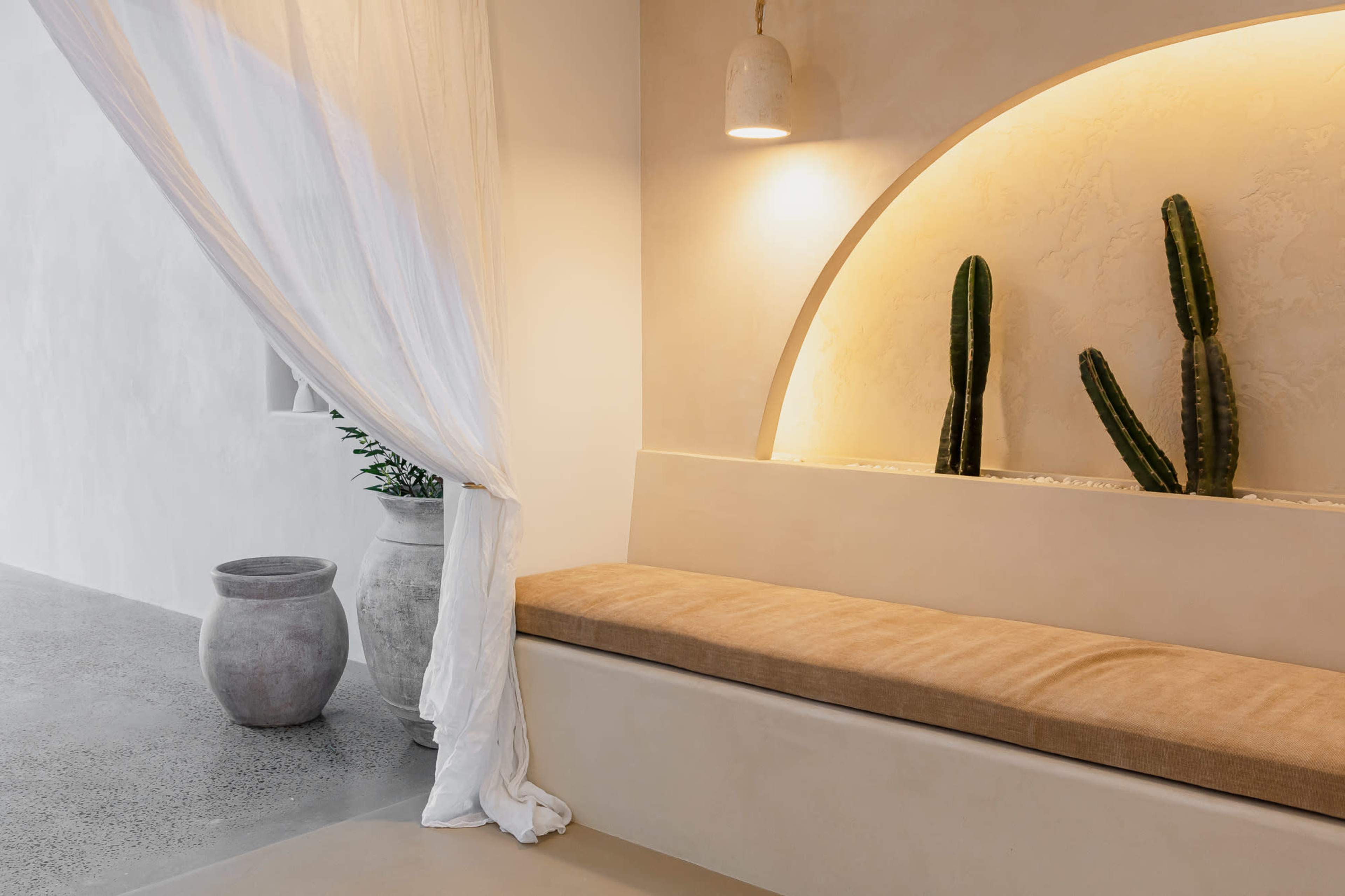 A minimalist indoor space features a beige bench with a soft cushion, illuminated by a wall light, and adorned with two tall cacti placed in an arched niche.