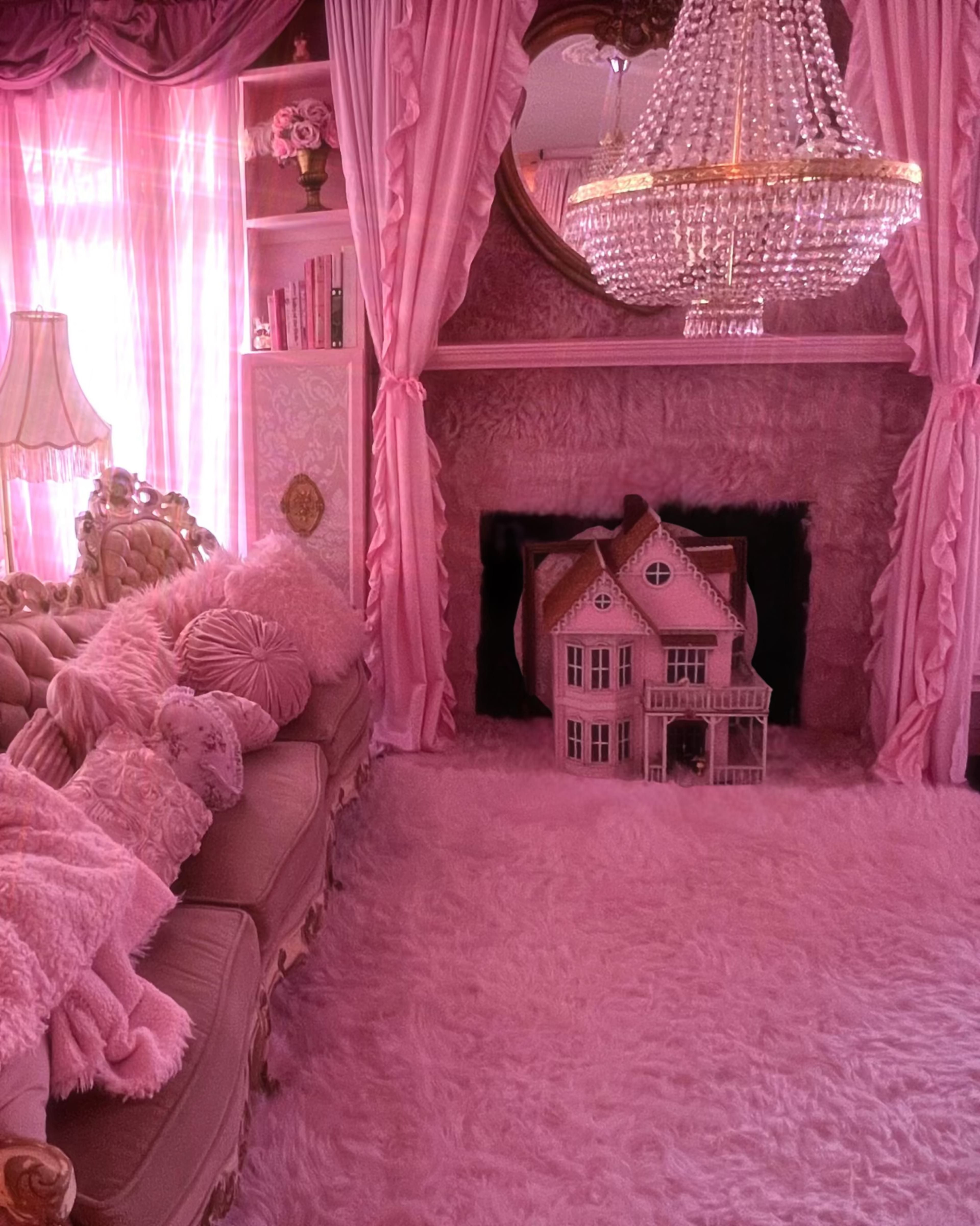 The room features a pink-themed decor with a plush rug, a chandelier, and a dollhouse positioned in front of a fireplace.