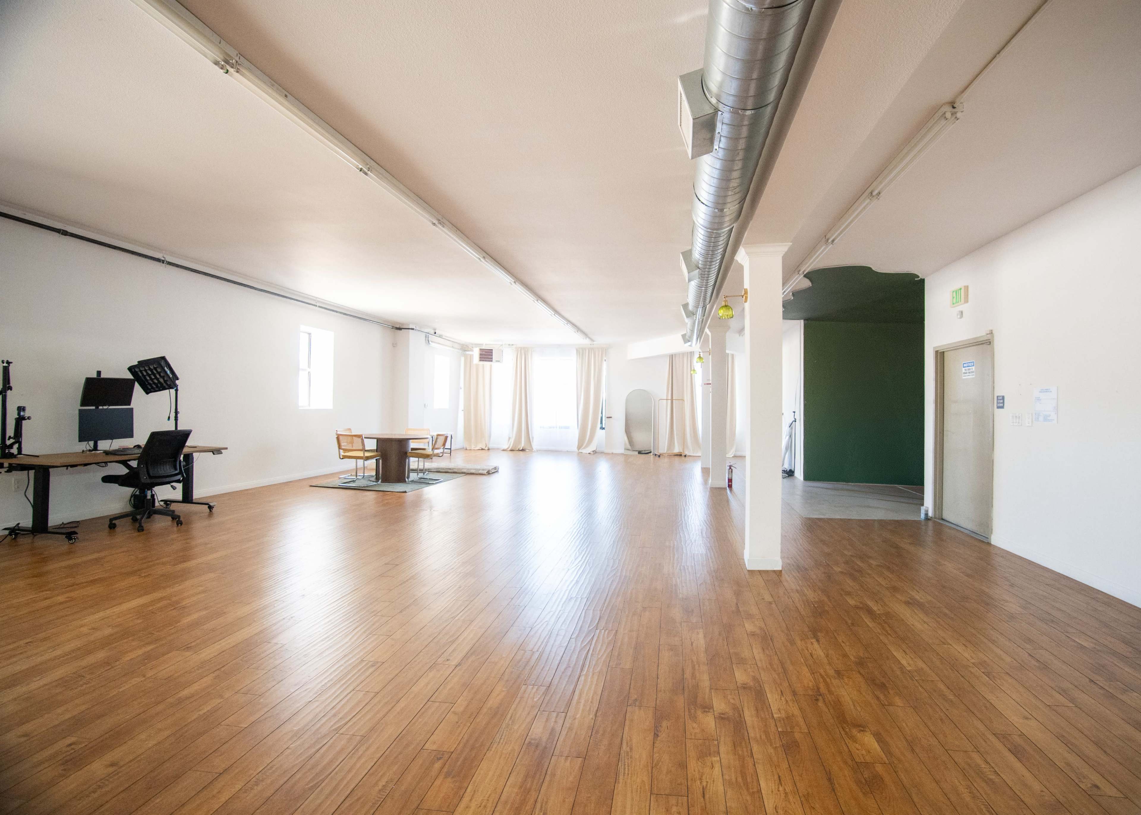 The image depicts a spacious, empty room with wooden flooring, featuring a dining table and chairs, a music stand, and an adjacent green wall.