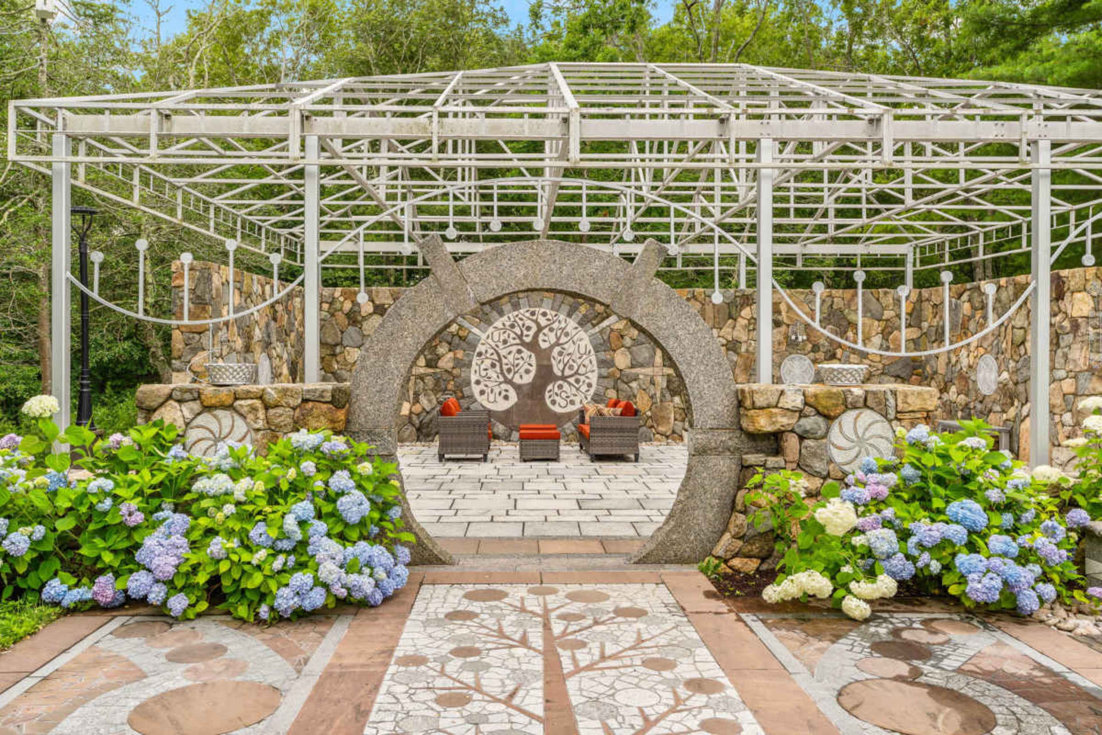 The image shows a landscaped garden area featuring a stone structure with circular designs and seating arranged among blooming hydrangeas.