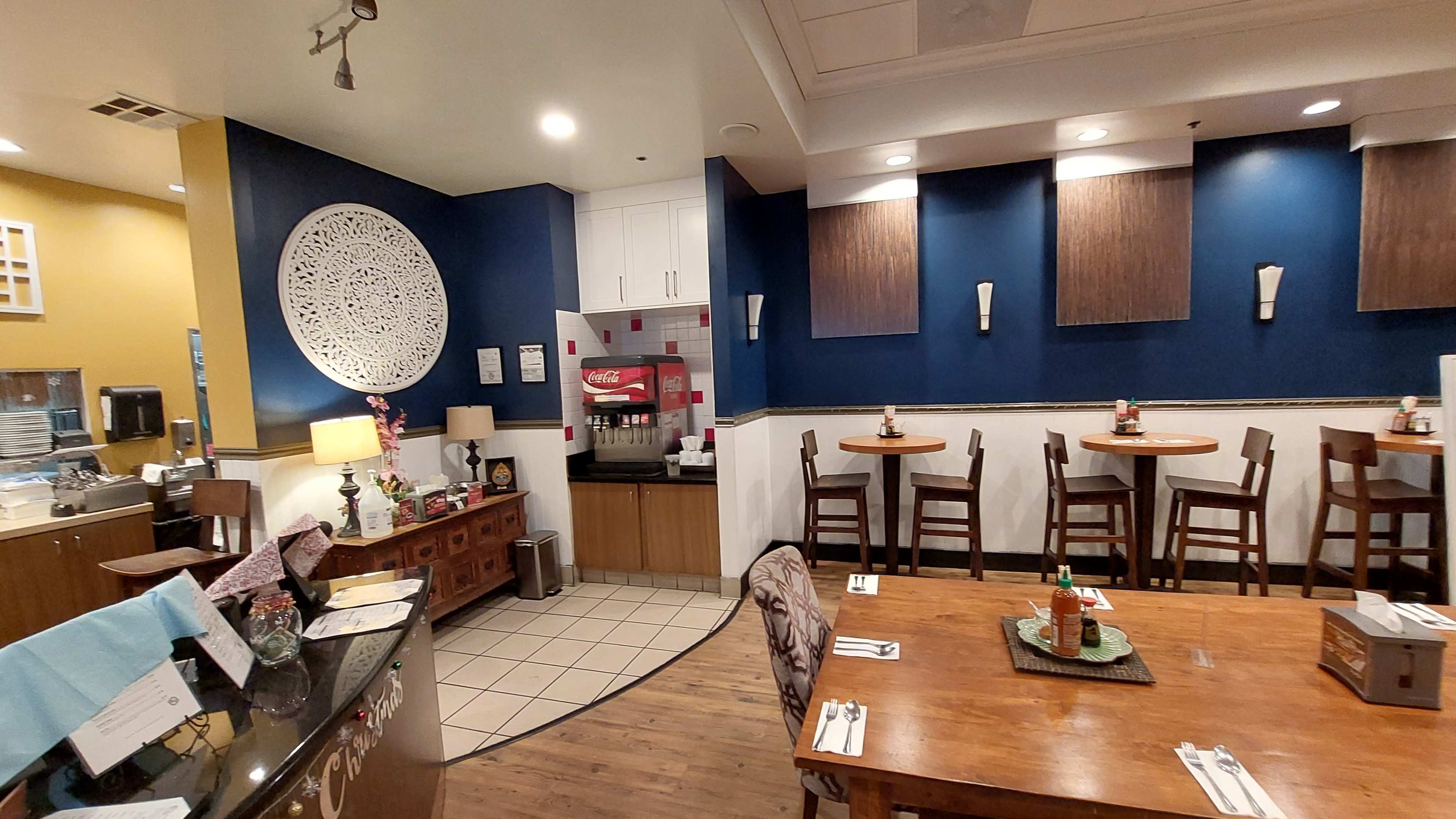 The image shows a restaurant interior featuring a mix of tables and chairs, a decorative wall design, and a beverage station.