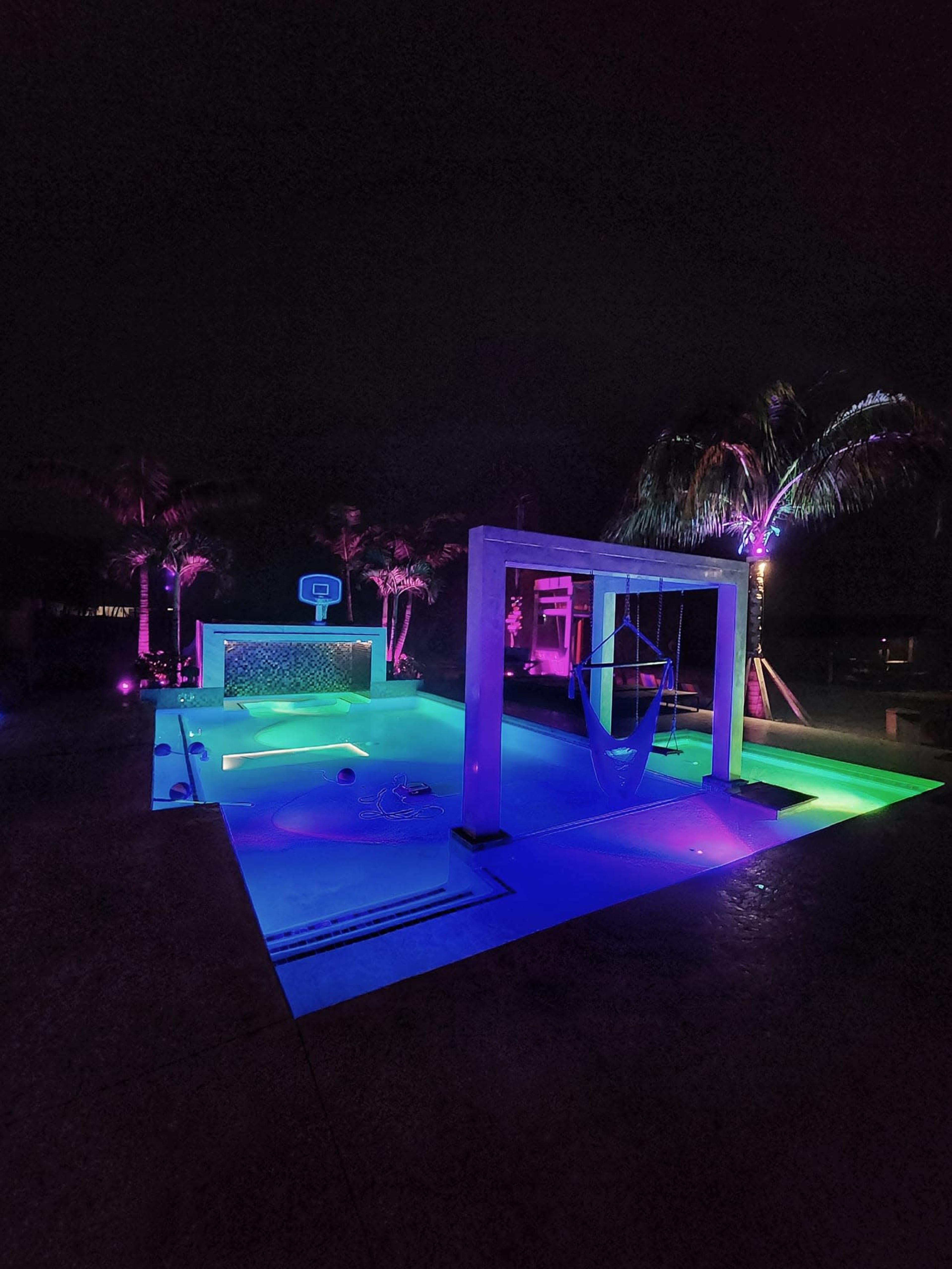 A brightly lit swimming pool area features colorful lights, palm trees, and a basketball hoop at night.