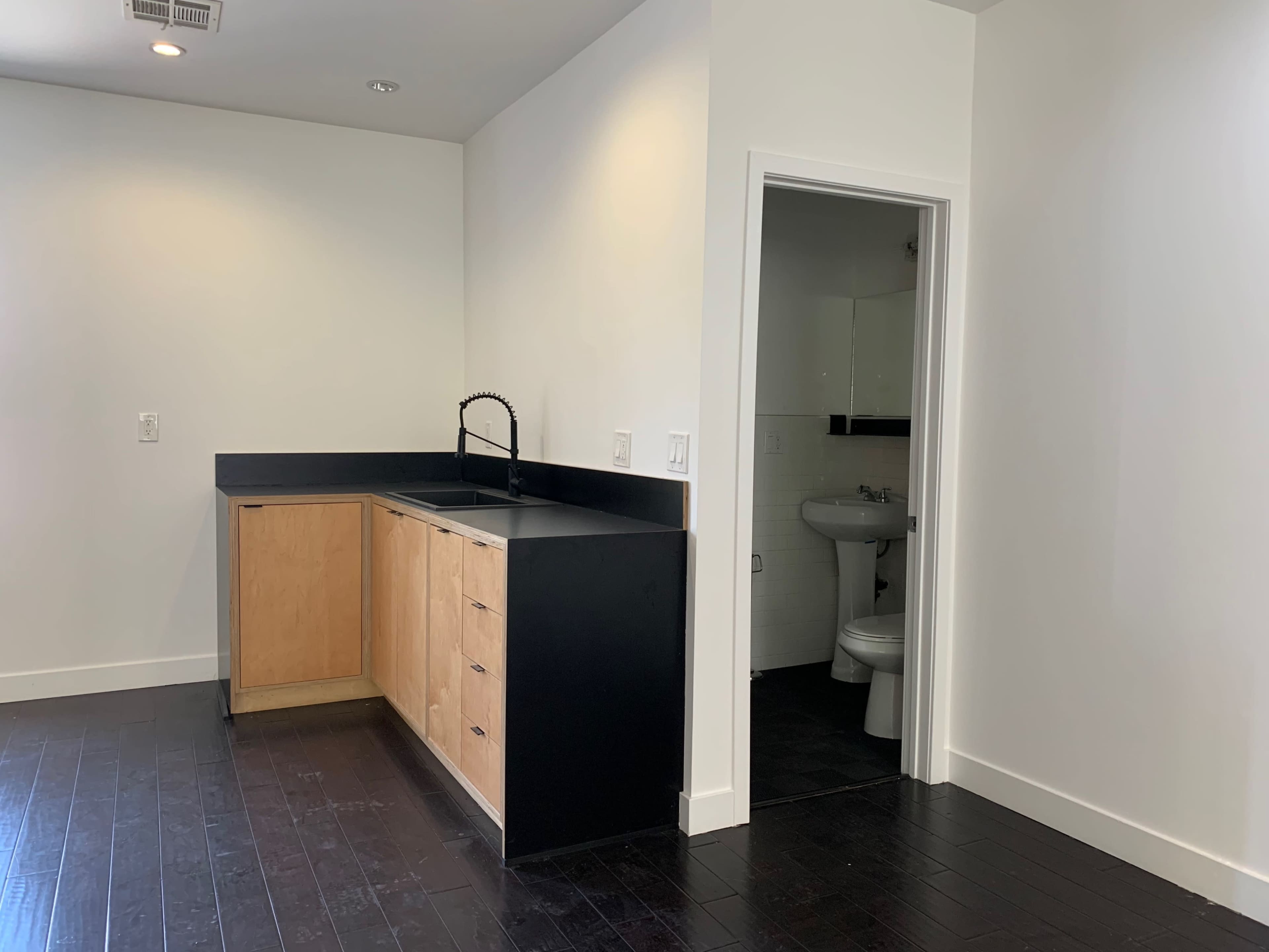 A small kitchen area with wooden cabinets and a black countertop adjacent to a bathroom doorway.