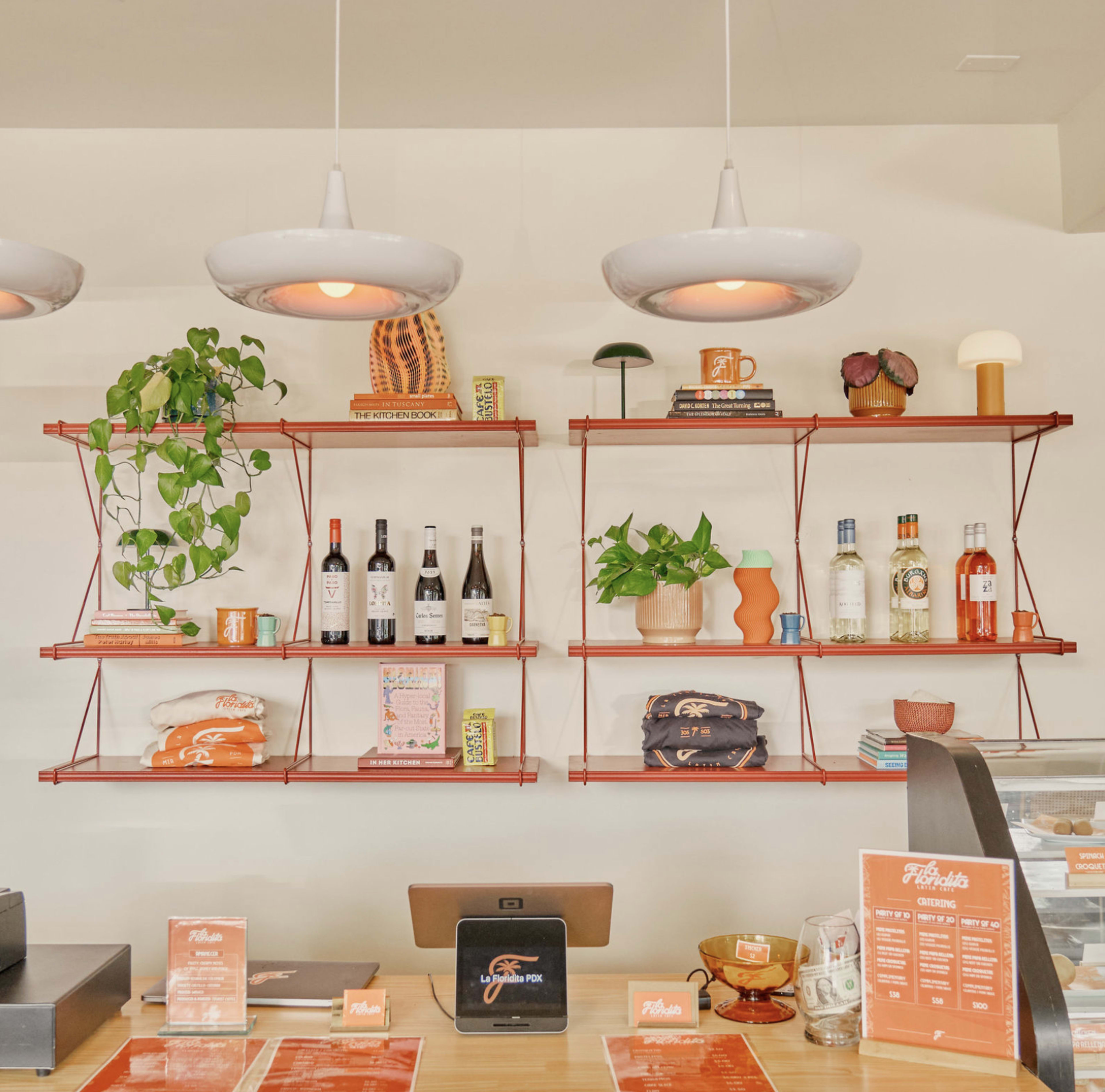 The image shows a modern café interior featuring two red metal shelving units filled with plants, books, bottles, and decorative items, along with a wooden counter displaying menus.
