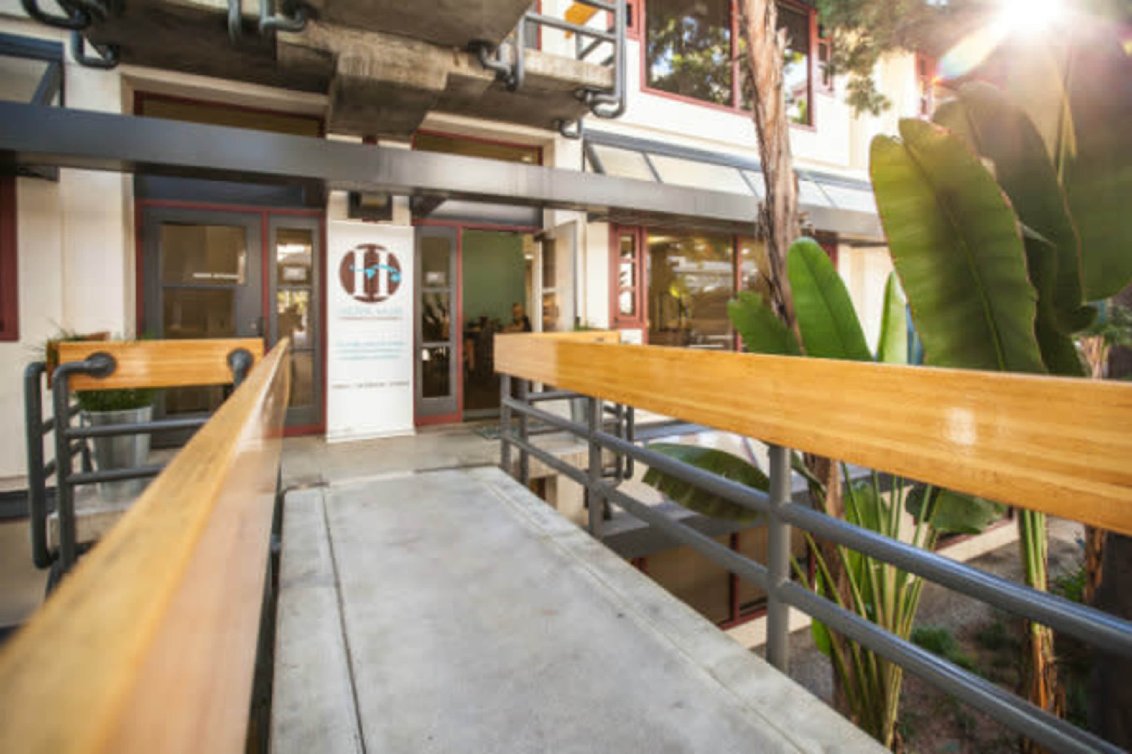 The image shows a concrete ramp leading to a building entrance, flanked by wooden handrails and tropical plants.
