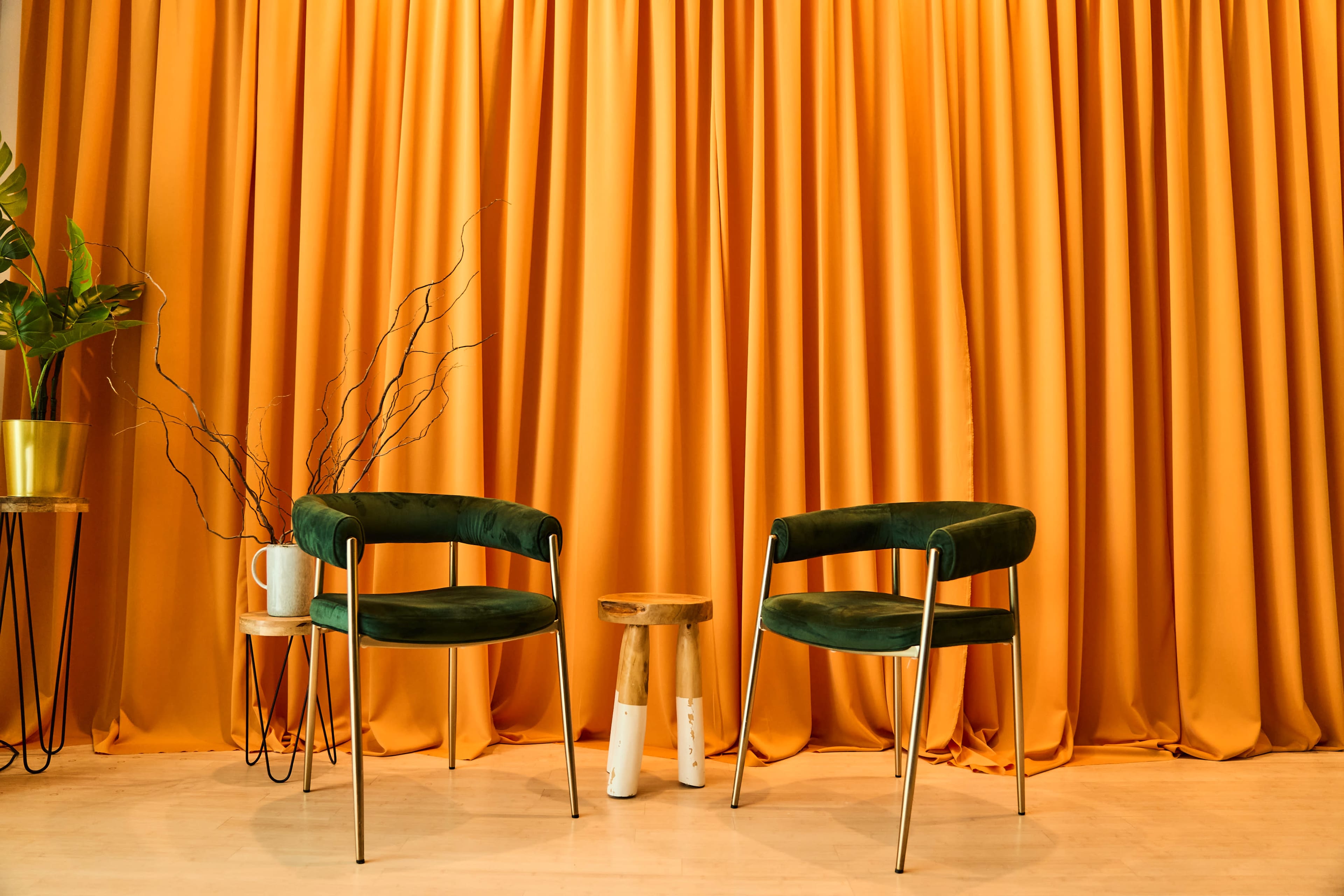 The image features two green velvet chairs positioned in front of an orange curtain, accompanied by a small wooden stool and a potted plant.
