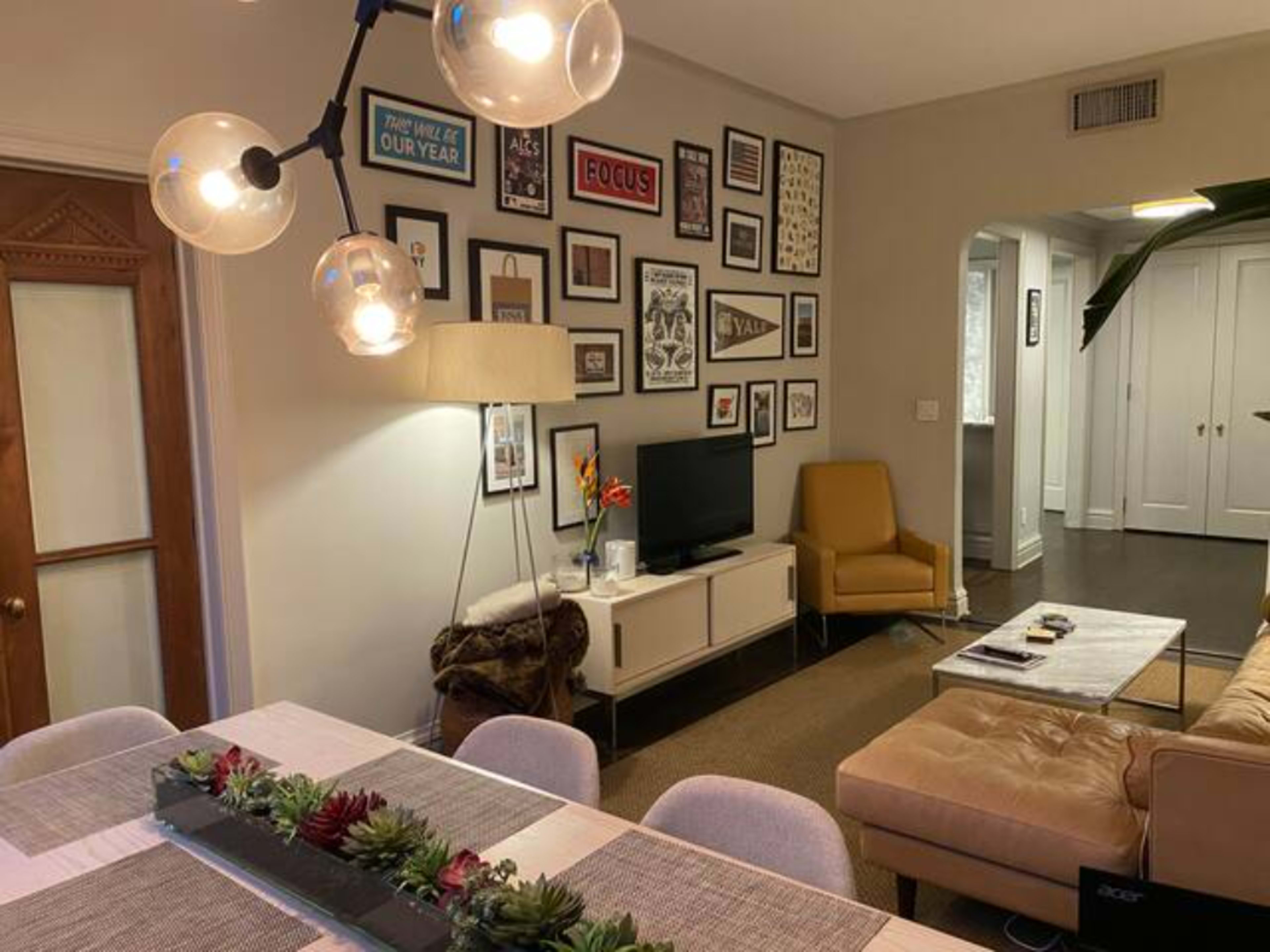 A living room features a television on a white cabinet, an orange armchair, a beige sofa, a dining table with a decorative centerpiece, and a wall decorated with various framed pictures.