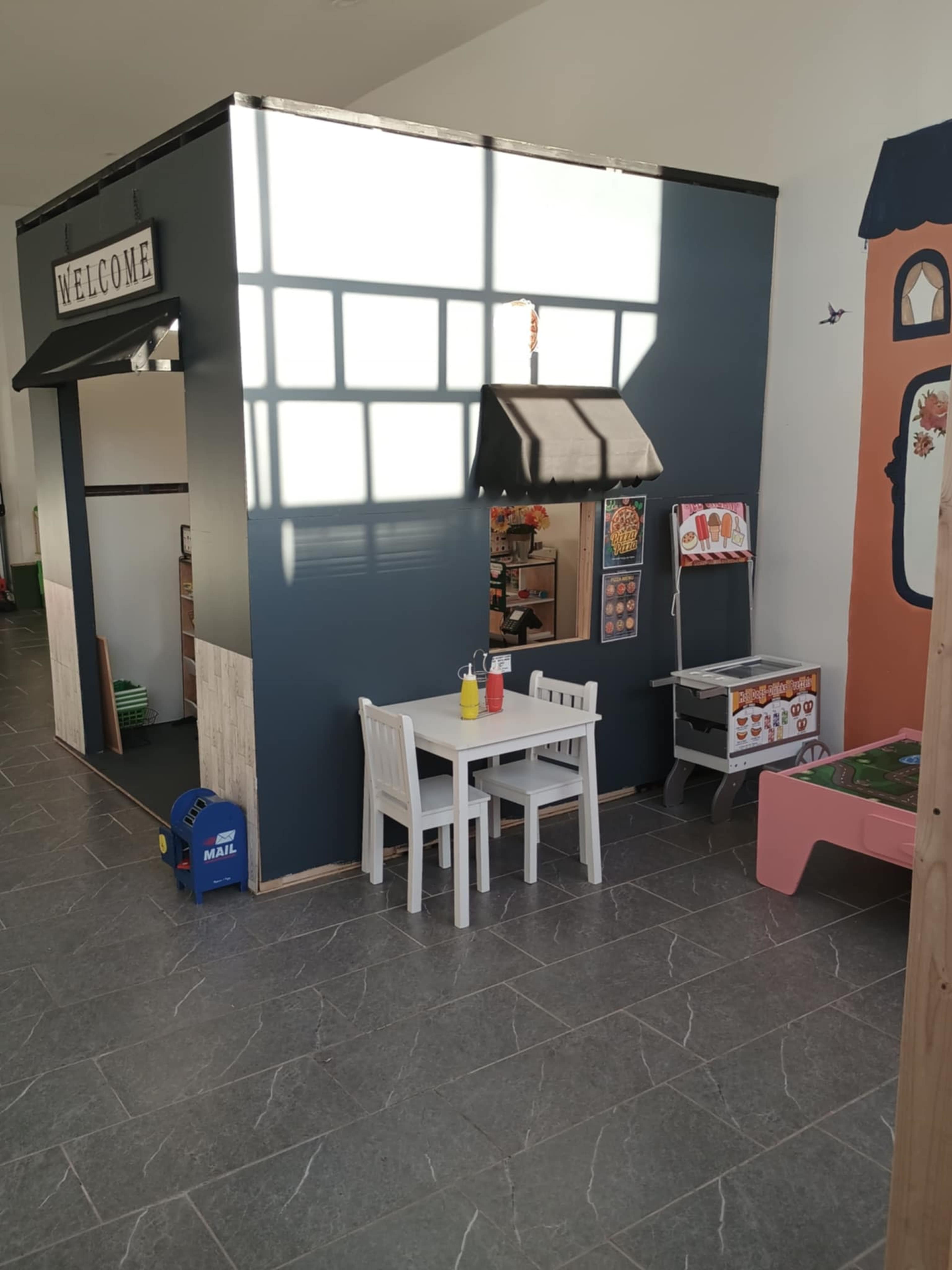 A small play area features a booth labeled "WELCOME," a tabletop with two chairs, and a cart displaying various toys and food items.