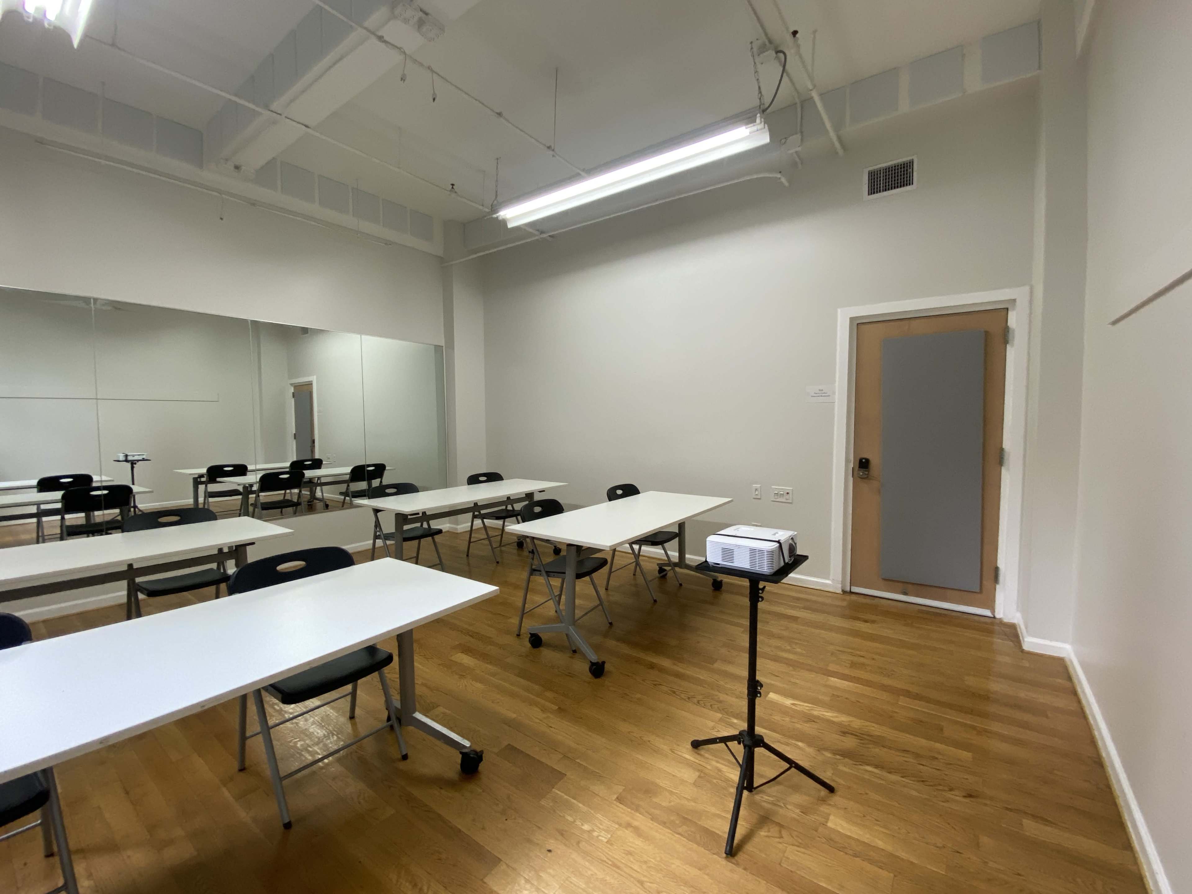 A vacant classroom with several white tables and chairs, a projector on a stand, and a large mirror on one wall.