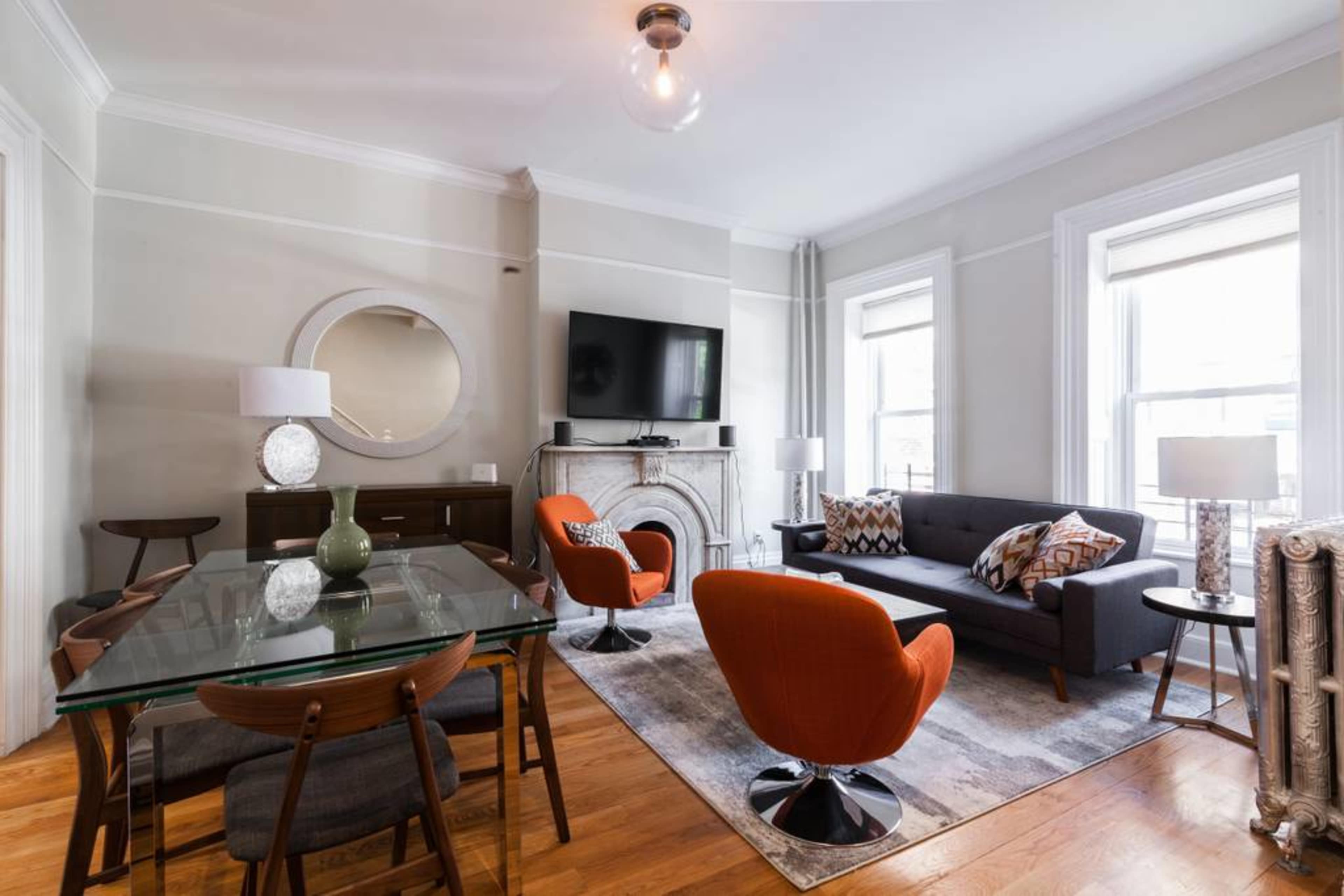 A modern living room with a glass dining table, two orange chairs, a gray sofa, and a marble fireplace, all illuminated by natural light from the windows.