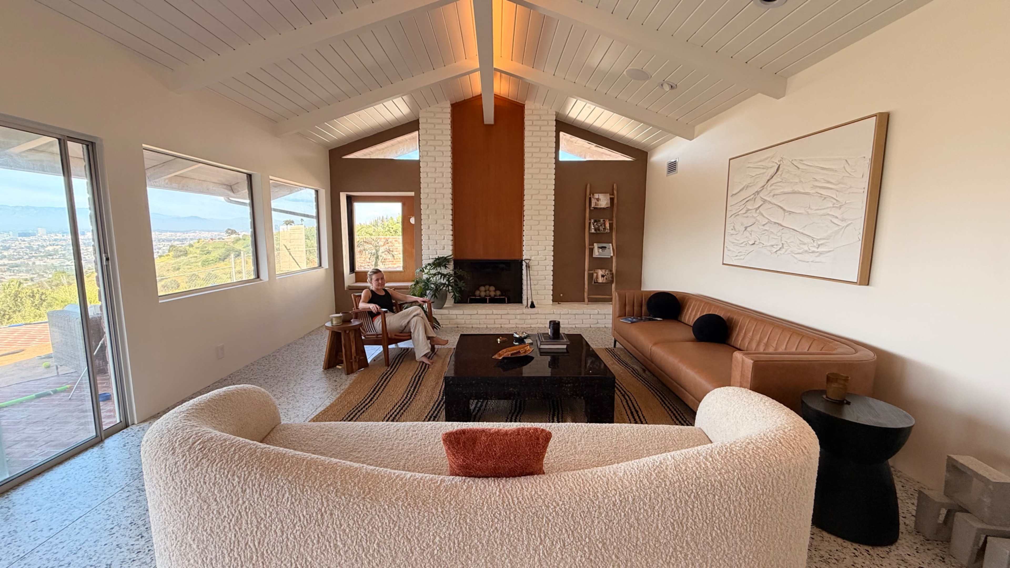 The image shows a modern living room with large windows, a fireplace, and a mix of contemporary furniture, including a tan leather sofa and a curved white couch.