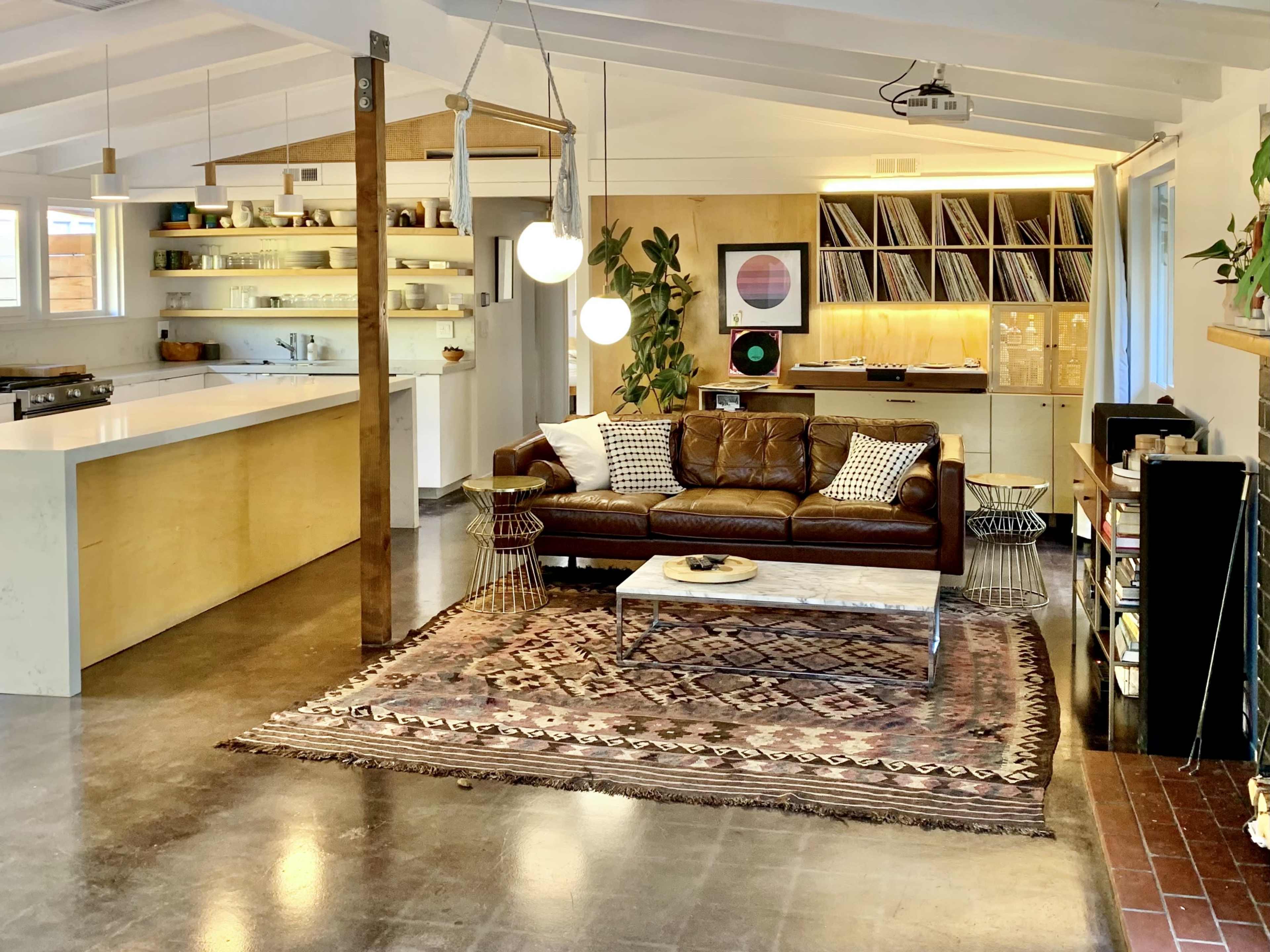 A modern indoor living space with a brown leather sofa, a large coffee table, an area rug, and a minimalist kitchen featuring open shelving and a wall of records.