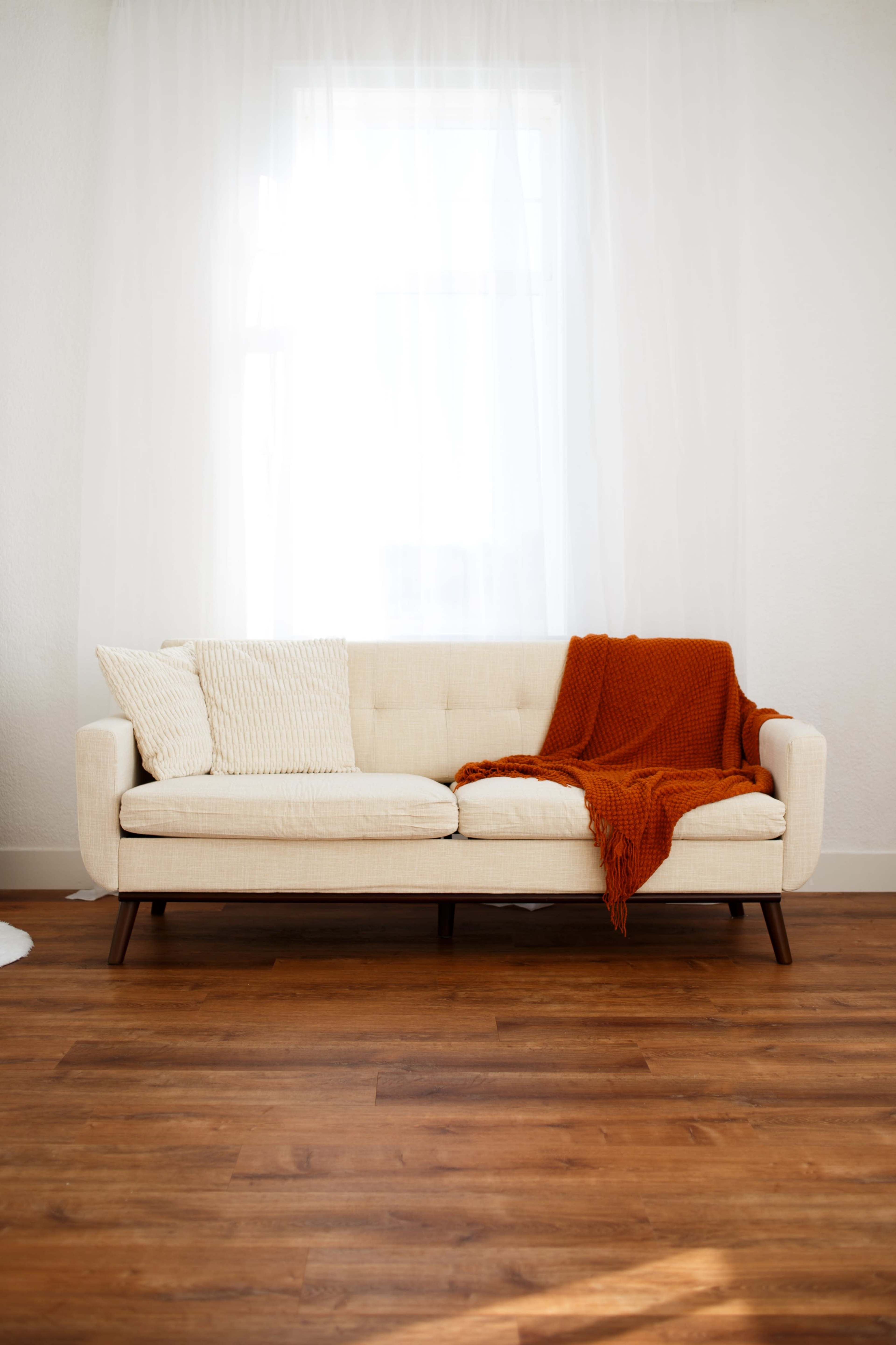 The image shows a cream-colored sofa with a textured white pillow and an orange throw blanket draped over it, positioned on a wooden floor beneath a bright window covered with sheer curtains.