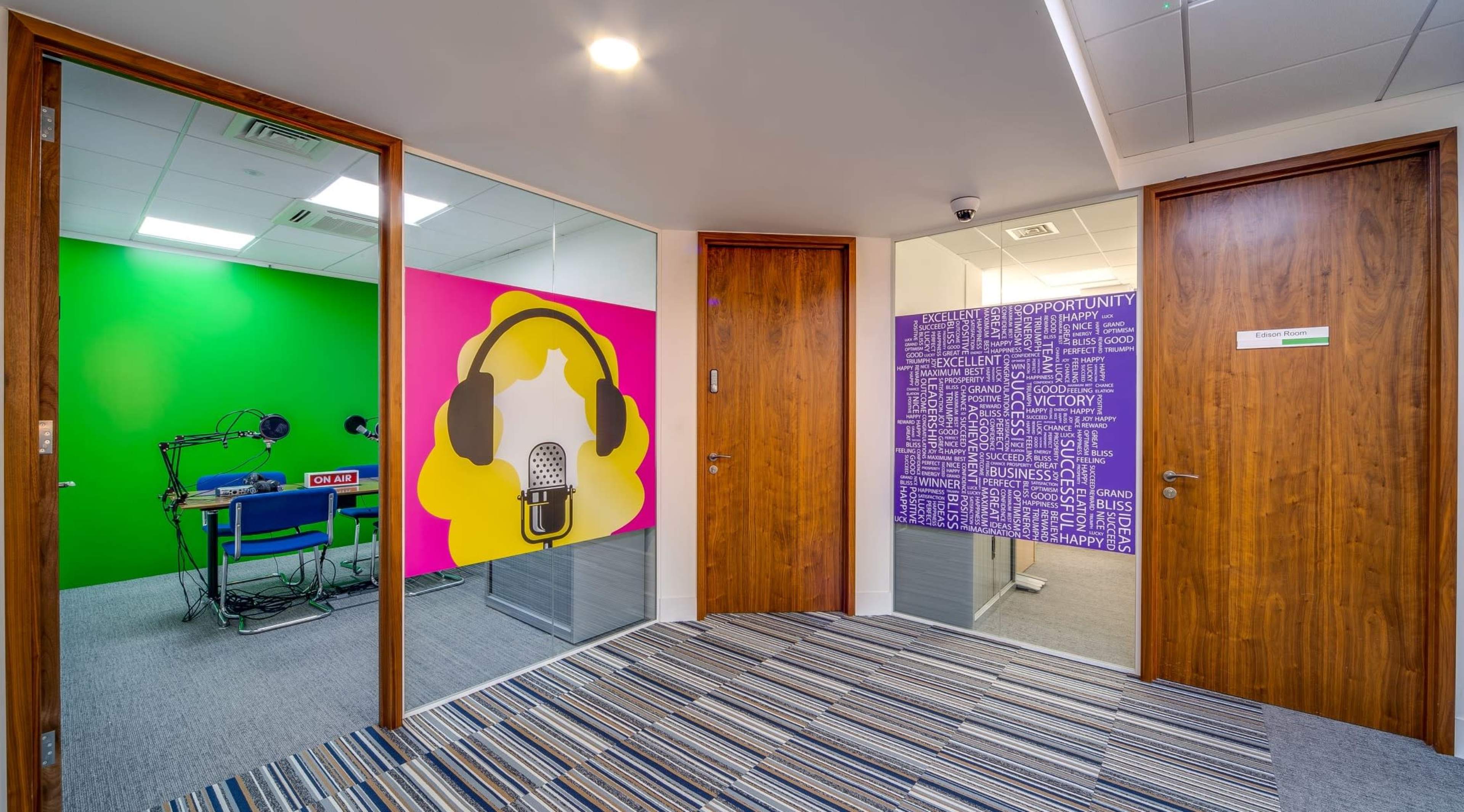 A modern office space featuring two glass-paneled rooms, one with a green wall and a microphone graphic, and the other with a purple wall covered in motivational words.