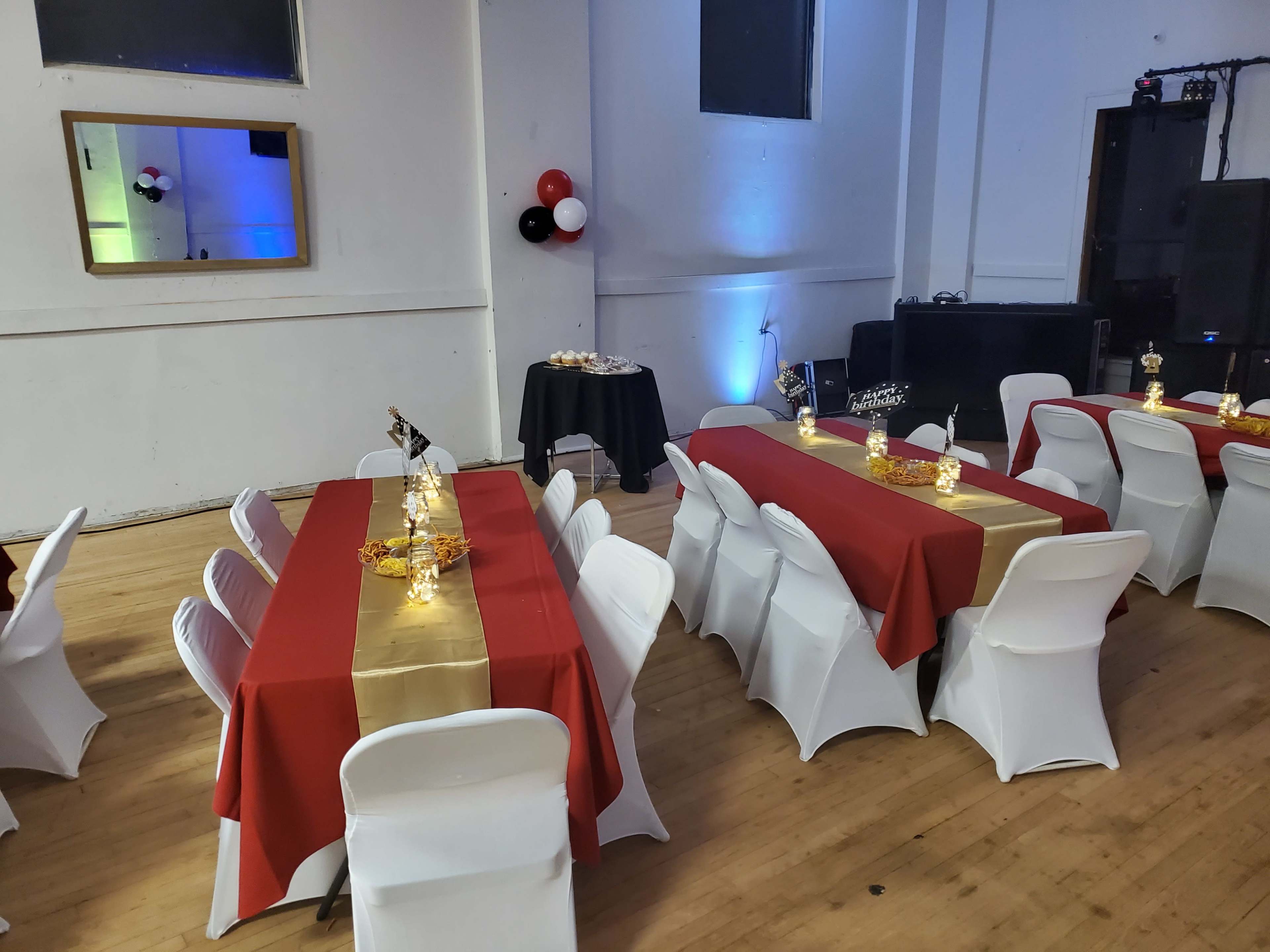 The image shows a banquet hall with neatly arranged tables covered in red and gold tablecloths, surrounded by white chairs, with decorative centerpieces and some balloons in the corner.
